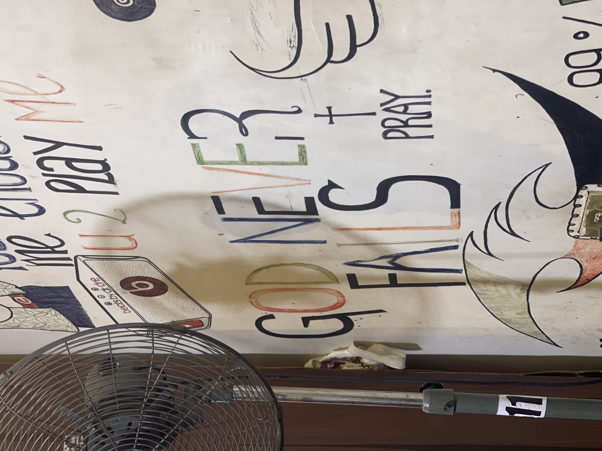The image shows a section of a wall with various handwritten words and drawings. In the foreground to the left, a metallic fan with a grey cage is visible. The fan is positioned in front of a wall. The wall itself is predominantly white and covered with colorful writing and illustrations.
Prominently displayed on the wall are several phrases:
- "GOD NEVER FALLS" is written in large, bold letters. The letters "GOD" are colored with red, orange, and green. The letters "NEVER" are in black and orange, and "FALLS" is in black with some red coloring on the 'A'.
- Above this, part of another phrase is visible, including "U2 PLAY ME". "U2" is in red, and "PLAY ME" is in black and red.
- Below "GOD NEVER FALLS", the word "PRAY" is written in black letters, with a small black cross above it.
To the left, there is a drawing of a white box with a red circular logo, labeled "beats by dr dre".
Further down on the wall, there are cartoon-like drawings, including what appears to be a black, pointed shape with wings or feathers above it. To the far right, a circular object with a spiral design is visible.
On the stand of the fan, a white sticker with the number "11" in black is attached.
The environment appears to be indoors, with a wall as the primary background. The writing and drawings suggest a creative or personal space, possibly a room or a community area. The presence of the fan indicates a warm climate. The overall impression is of a wall that has been used as a canvas for expressive art and messages.