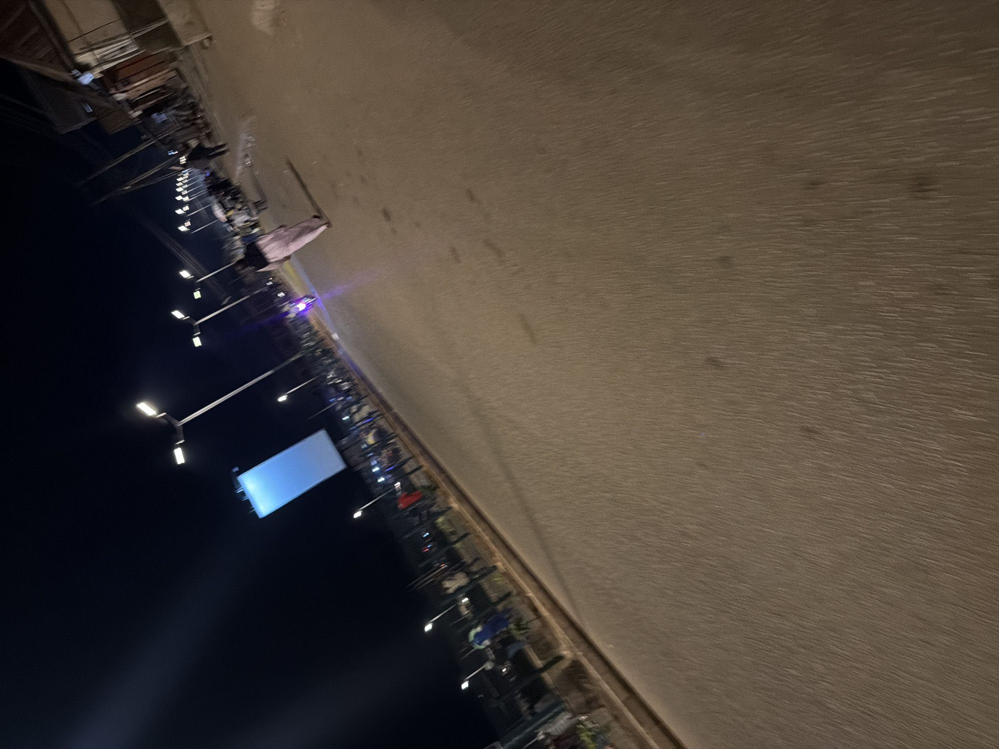 This is a nighttime, horizontal, low-angle, slightly blurred photograph taken in Bukuru, Nigeria. The scene appears to be an outdoor market or street lined with stalls. In the foreground, illuminated by streetlights, are various objects, possibly merchandise, displayed under awnings and tables. Some of these objects are covered with fabric, giving them a soft, indistinct appearance. Several individuals are visible, though their features are unclear due to the low light and motion blur. They appear to be interacting with the stalls or walking through the area.
The background is dominated by a dark, unlit sky on the left and a textured, concrete-like surface on the right, possibly a wall or the ground, illuminated by overhead lights. There are multiple lampposts with bright lights spaced along the scene, casting light on the path and the stalls. One lamppost in the mid-ground has a large, illuminated rectangular sign attached to it, displaying a bright blue light. The overall impression is that of a bustling, active marketplace at night. The blurred motion suggests that the photograph was taken while moving.