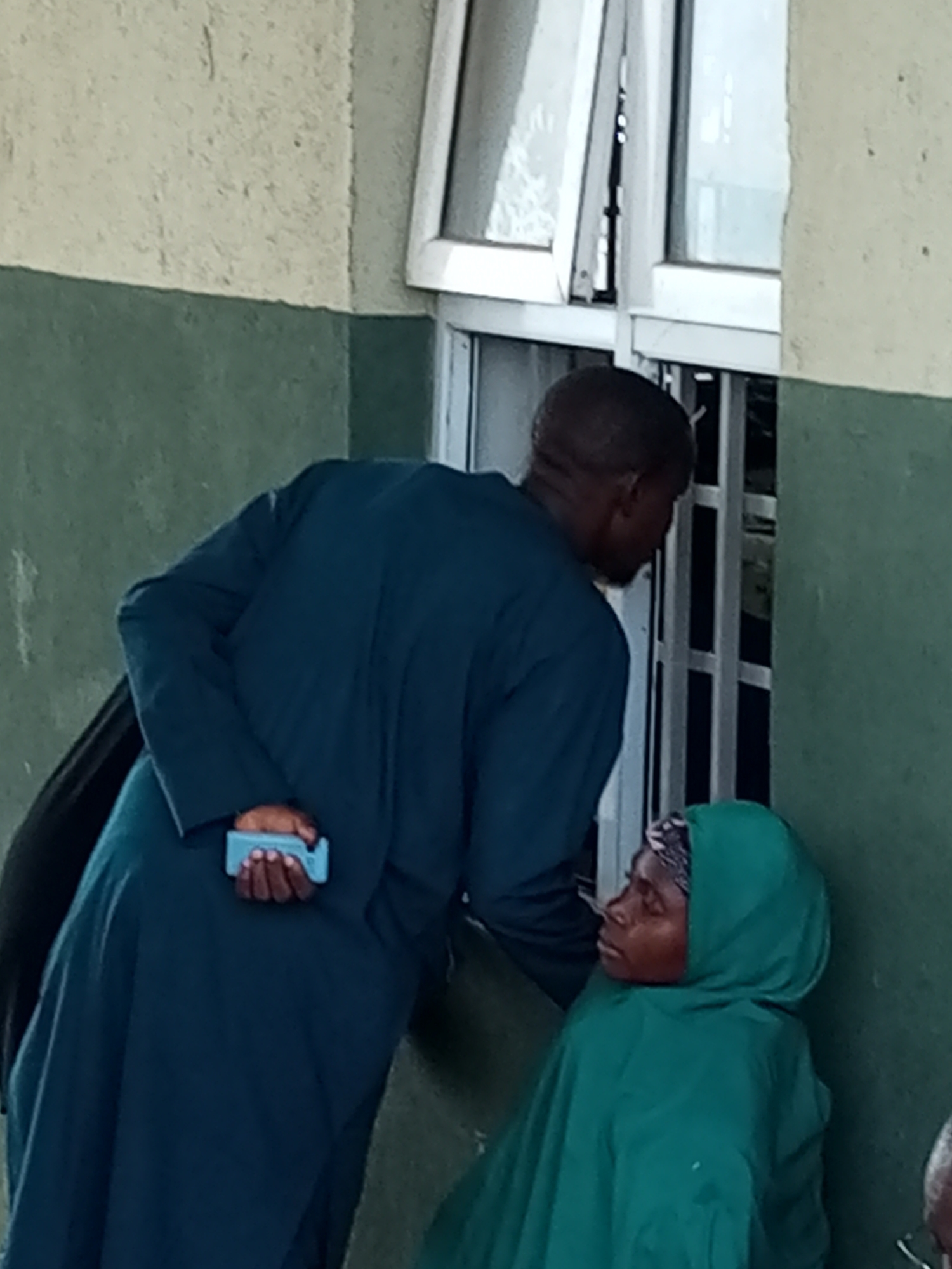 In Gombe, Nigeria, a man wearing a dark blue robe leans towards a seated woman who is covered in a green hijab. The man holds a small, light blue object in his right hand. They are positioned next to a building with a green and beige exterior, and a window with white frames. The scene appears to be outdoors or in a covered walkway.