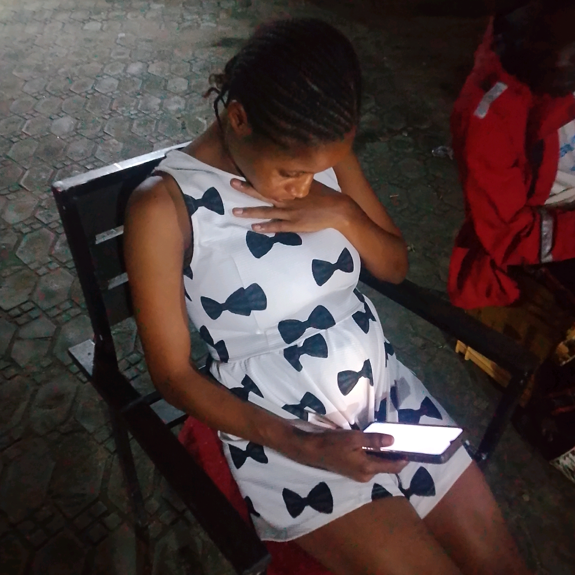 A pregnant woman wearing a white dress with black bow patterns sits in a dark, outdoor setting with a cobblestone floor. She is holding a smartphone and looking down at it. The scene appears to be in Ado, Nigeria.