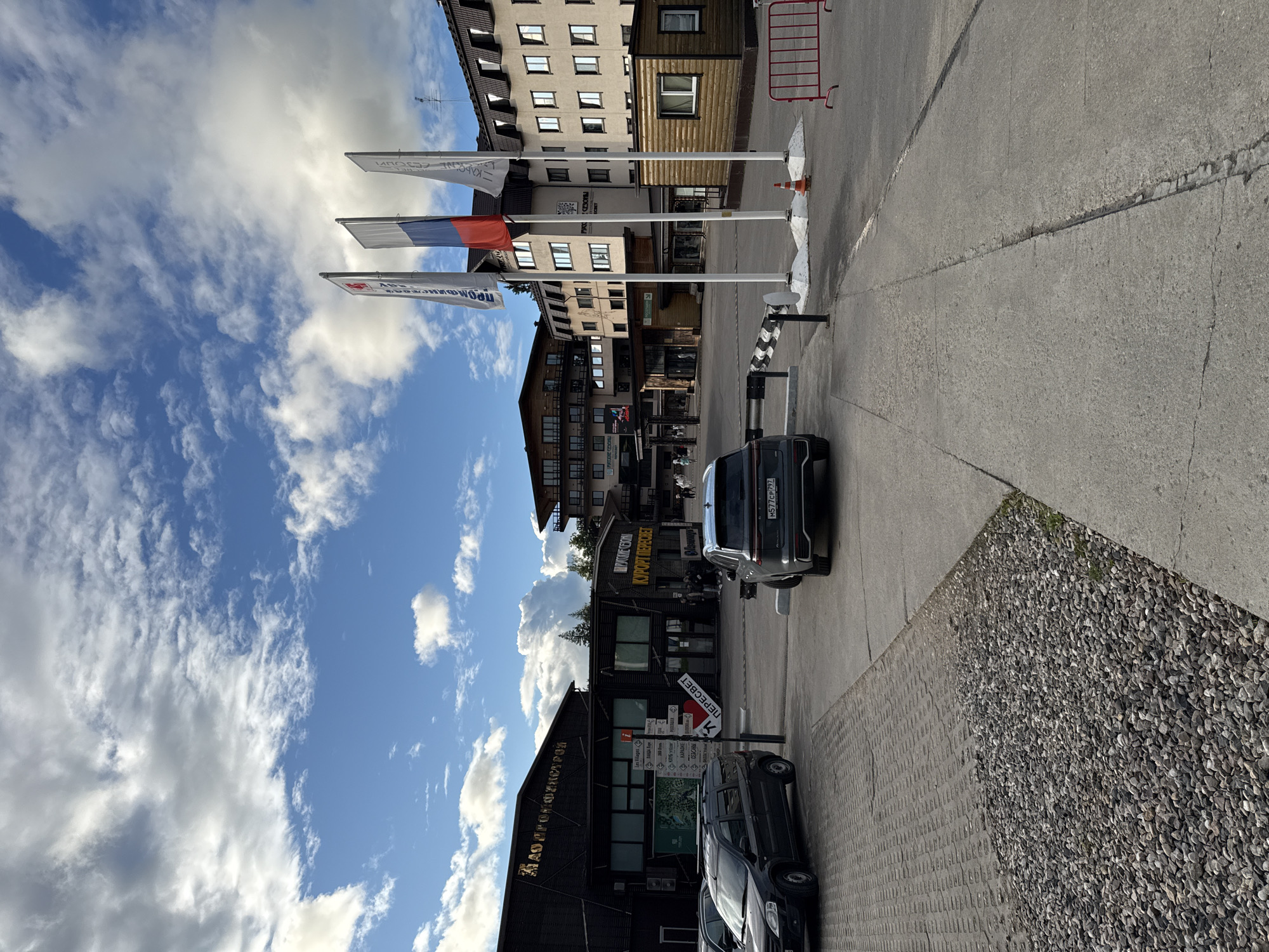 The image captures a street scene in Peresvet, Russia. In the background, there are buildings with multiple floors, and flags. The middle flag is the Russian flag. A dark-colored car is parked along the street. A sign that includes the text "АО ПРОЙФИНСТУРОЙ" is on a building. Another sign reads "КУРОРТ ПЕРЕСВЕТ". The sky is blue with scattered clouds. The street is paved with concrete and a section is covered in gravel. A sign points directions to "Les Villages", "КАРАОКЕ", "СЕТОНЫЫ", "ПЕРЕСВЕТ", "РУССКИЕ СЕЗОНЫ" (the text on the sign may be partially obscured). The license plate of the car in the front says "M577CP 797".