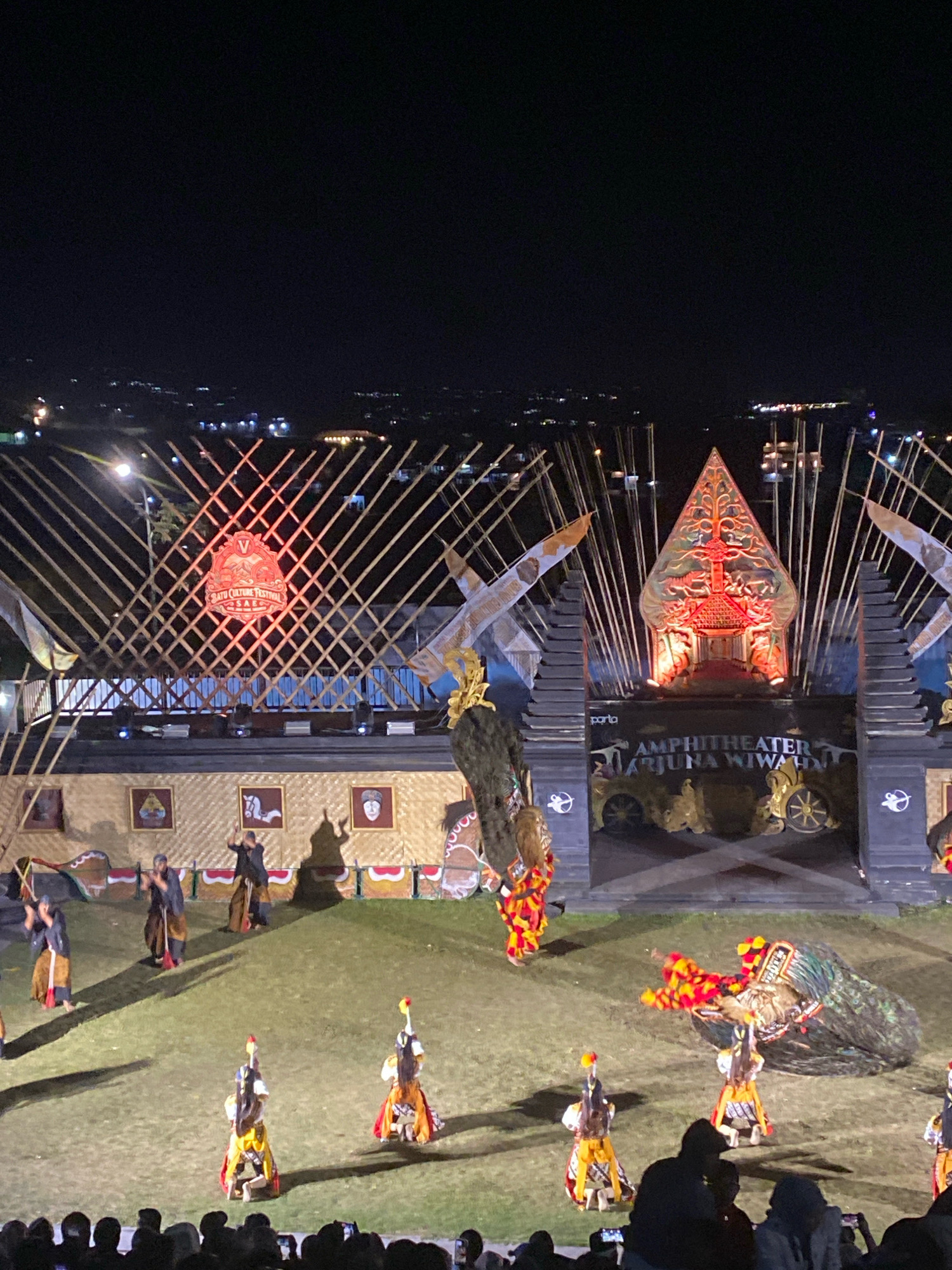 An outdoor amphitheater stage is illuminated at night for a cultural performance. The stage features elaborate decorations, including bamboo structures, traditional Javanese shadow puppet motifs (Gunungan), and a sign that reads "SATU CULTURE FESTIVAL" and "AMPHITHEATER ARJUNA WIWAHA". A group of performers in colorful traditional costumes are on the grassy stage, some engaged in dance or ritualistic movements. In the foreground, a large, peacock-like costume is prominent, and in the background, the lights of a city are visible on a hill. An audience is seated in the foreground, with some holding up their phones to record the event.