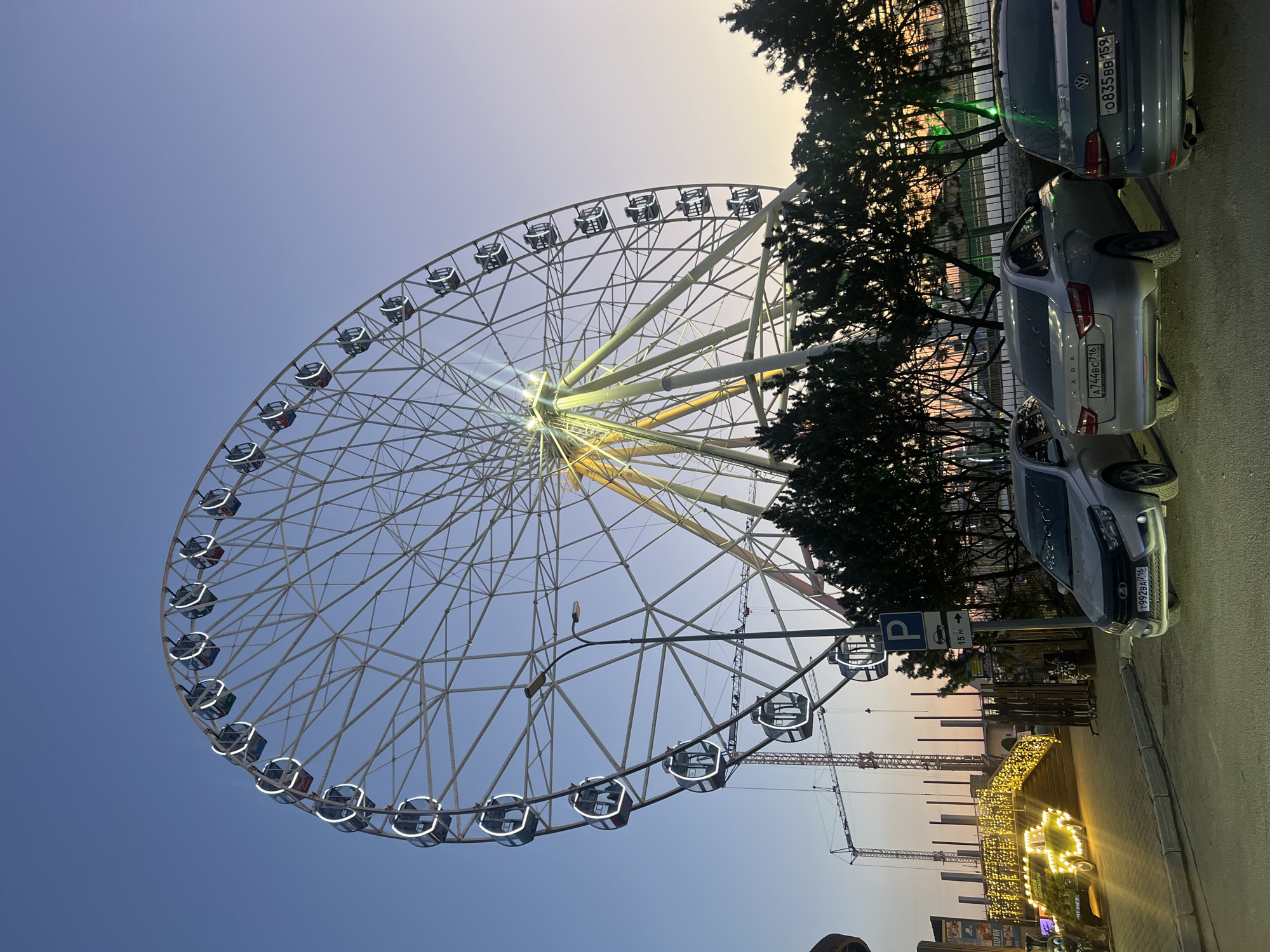 A large Ferris wheel with white frame, cabins, and lights dominates the view against a dusk sky, fading from blue to orange near the horizon. The wheel is positioned to the left, partially obstructing the view of trees and parked cars on the right side. At the bottom, there are decorations with lights, resembling an illuminated archway. Above the decorations are construction cranes and a parking sign. The license plate of a parked Lada car in Russian is visible - A744BC716. The location is likely in or near Kazan, Russia.