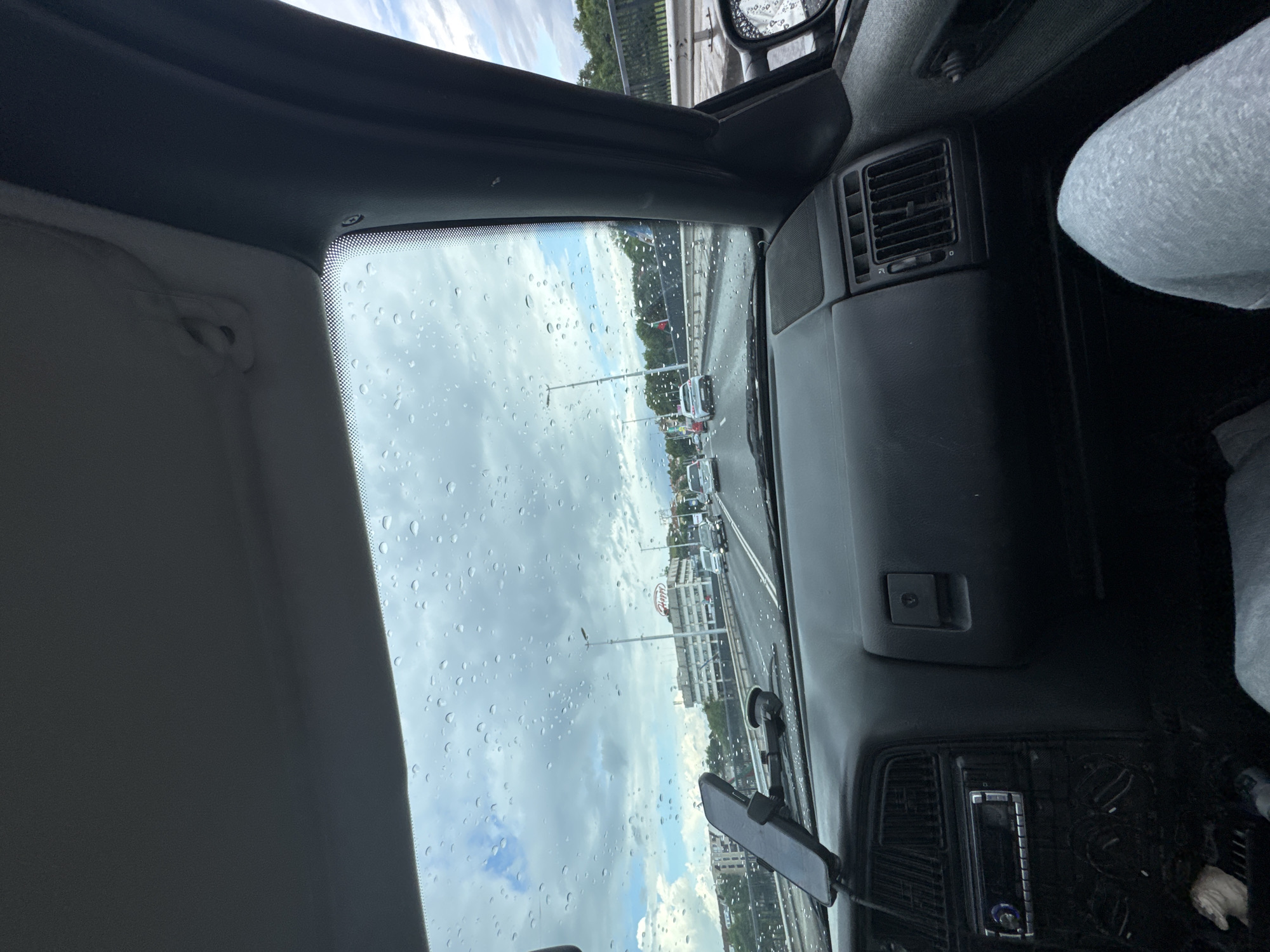 A passenger is sitting in the front seat of a vehicle, likely on a bridge in Plovdiv, Bulgaria, as indicated by the Happy Bar & Grill restaurant visible through the windshield. Raindrops cover the windshield, partially obscuring the view of traffic and buildings. The passenger's leg is visible, wearing light-colored pants. A phone is mounted on the dashboard. The vehicle's interior is gray and includes air vents and a radio. Other vehicles are on the road. The sky is cloudy.