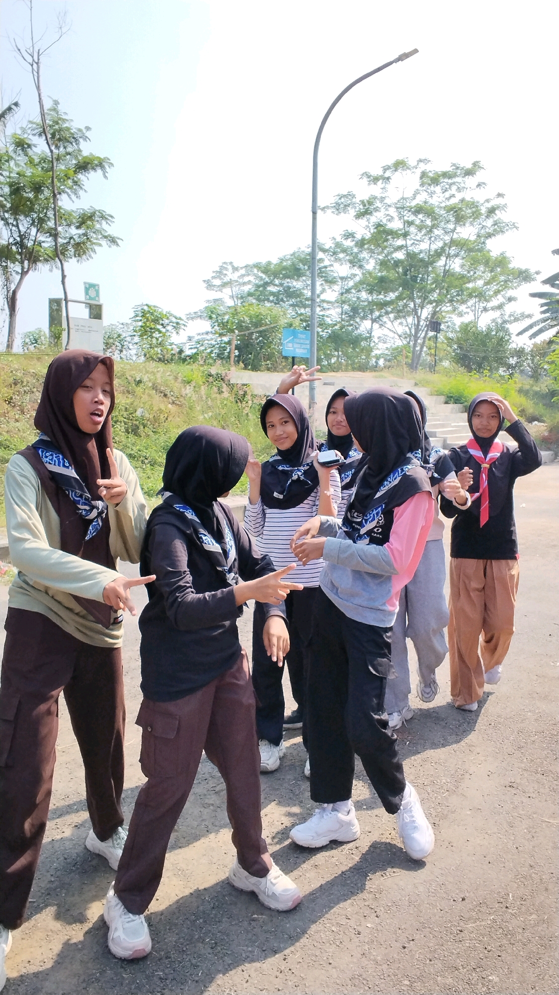 The image shows a group of six young females, likely students, posing for a photograph outdoors. They are all wearing headscarves and uniforms, suggesting they are part of a scout or similar youth organization. The attire includes various colored hijabs and matching scarves with a blue and white pattern, worn over tops and trousers in shades of brown, black, grey, and pink. Three of the individuals are making peace signs with their fingers. The setting appears to be a paved path or road, with a grassy embankment and trees in the background. A street lamp is visible on the right side of the image, and some signs are present in the distance. The overall lighting indicates a bright, sunny day. The exact location is not evident from the visual cues, and therefore cannot be identified as Ngingasrembyong, Indonesia.