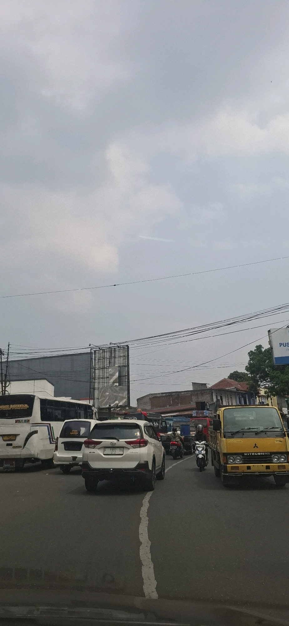 The image depicts a traffic jam on a city street in Sirnagalih, Indonesia. A white SUV with license plate F 1302 YH is in the foreground, facing away from the camera. To its left is a white bus and another white vehicle. A yellow Mitsubishi truck is visible on the right side of the road, also facing away. Several motorcycles are interspersed in the traffic, with one rider wearing a green jacket. The sky is overcast with scattered clouds. Overhead power lines are strung across the scene, and buildings are visible in the background. The road surface is asphalt with a white lane divider.