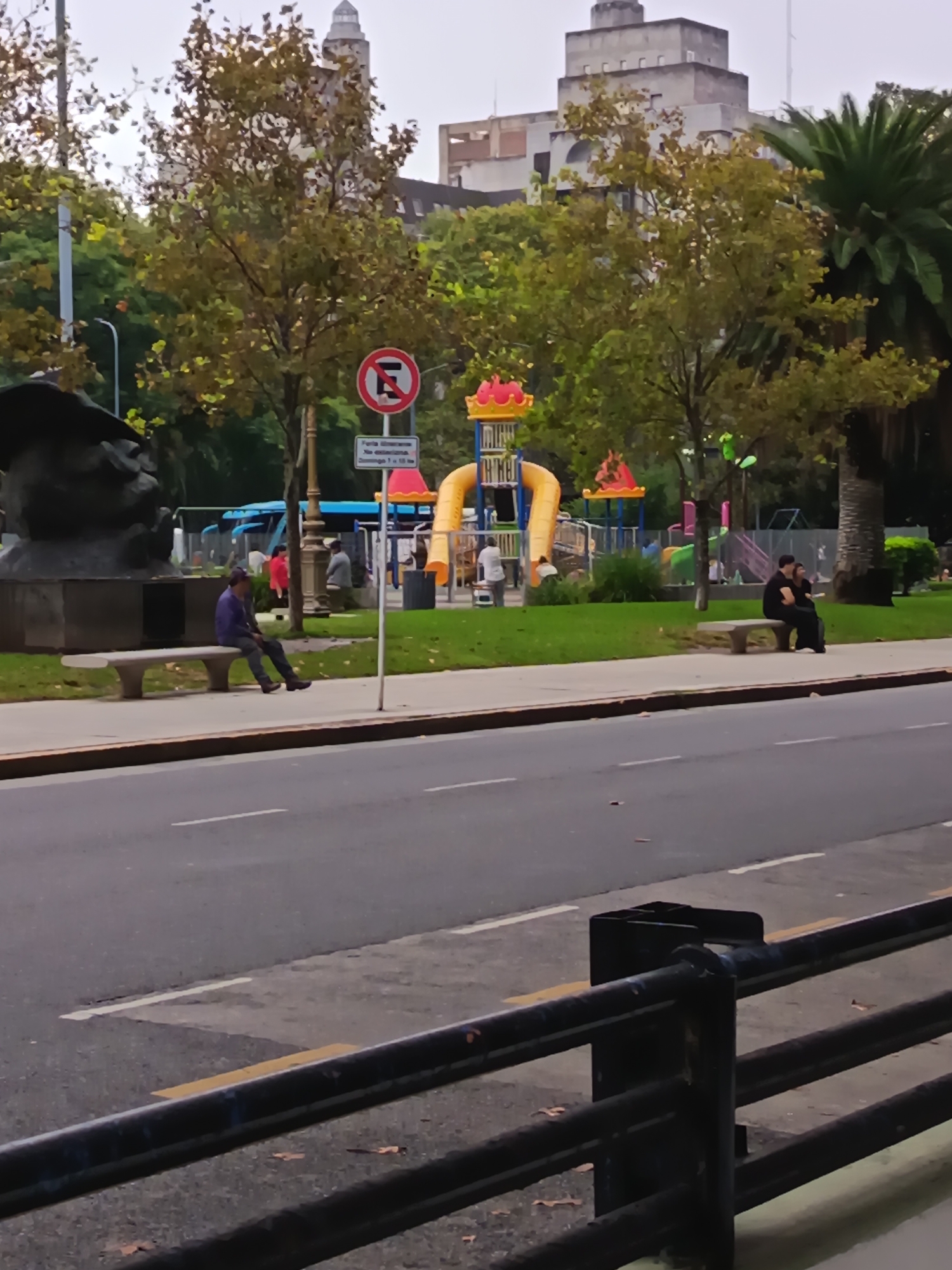 The image shows a street scene in Buenos Aires, Argentina. A wide, paved road separates the foreground from a park area in the background. In the foreground, a black metal railing is partially visible.
In the park area, there's a playground with colorful equipment. Several people are present: one person sits alone on a bench near a statue of a man with a bust, while a couple sits on another bench further in the park. Additional people are scattered throughout the park, some near the playground. A "No Parking" sign is visible near the benches and playground.
Behind the park, buildings are visible, including one with a tower. Trees with greenish-yellow foliage are dispersed throughout the park. The sky is overcast, indicating potentially cool weather.