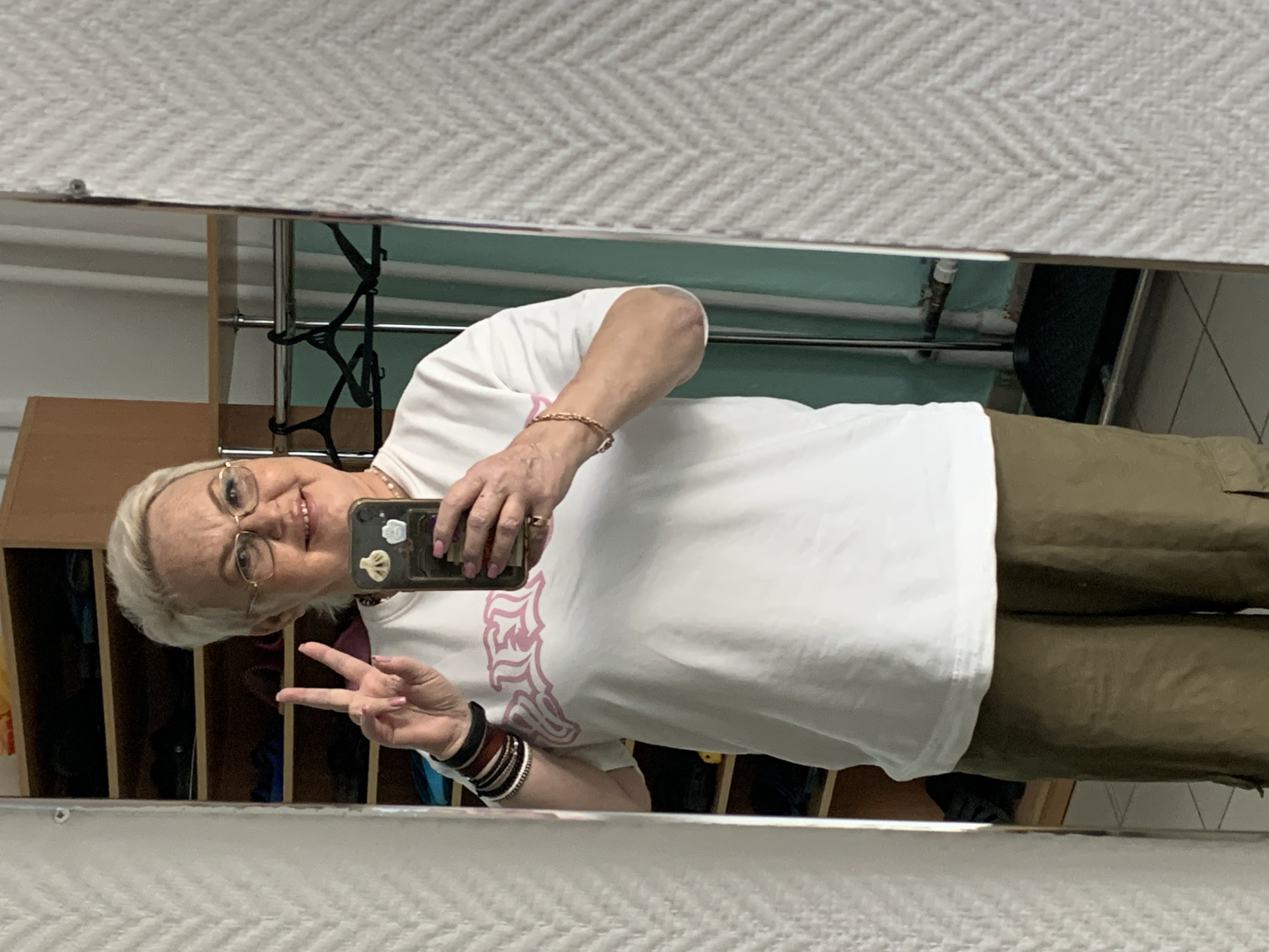A woman with short, blonde hair and glasses is taking a selfie in front of a mirror. She is wearing a white t-shirt with a pink design, olive green pants, and several bracelets on her left wrist. She is holding a smartphone with multiple stickers on the back and making a peace sign with her left hand. Behind her, in the reflection of the mirror, is a room with a light blue wall, a wooden cabinet, a clothing rack with hangers, and possibly other furniture. The location is Chaikovsky, Russia, based on the t-shirt.