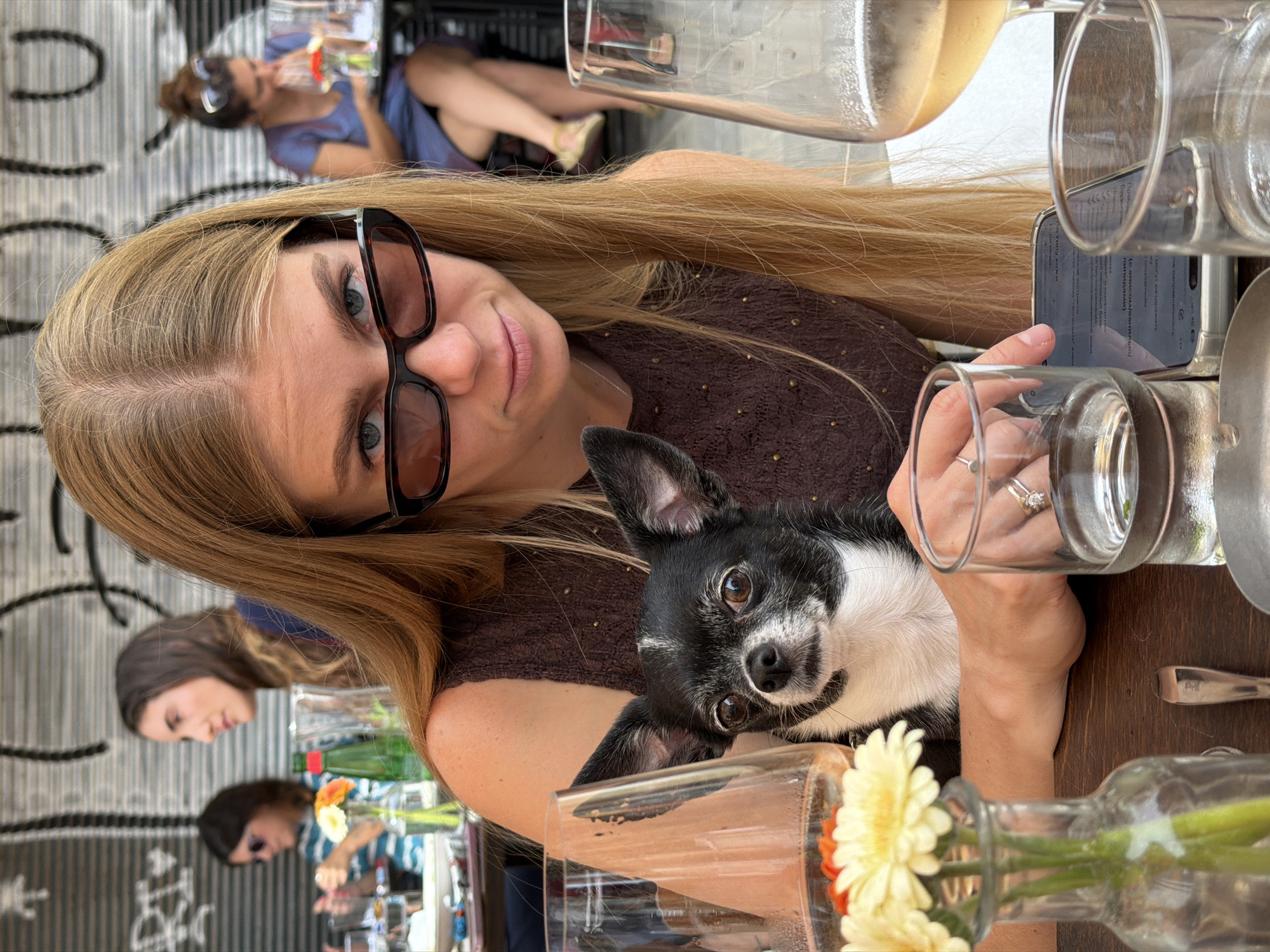 A woman with long blonde hair and large black-rimmed glasses holds a black and white Chihuahua in her arms at a table. Her hand is holding a clear glass. There is a mobile phone on the table next to the glass. Other clear glasses and vases with flowers are also on the table. Several other women are seated at tables in the background, some holding drinks. The picture was taken at a restaurant in Tel-Aviv, Israel. A wall in the background has a black and white design.