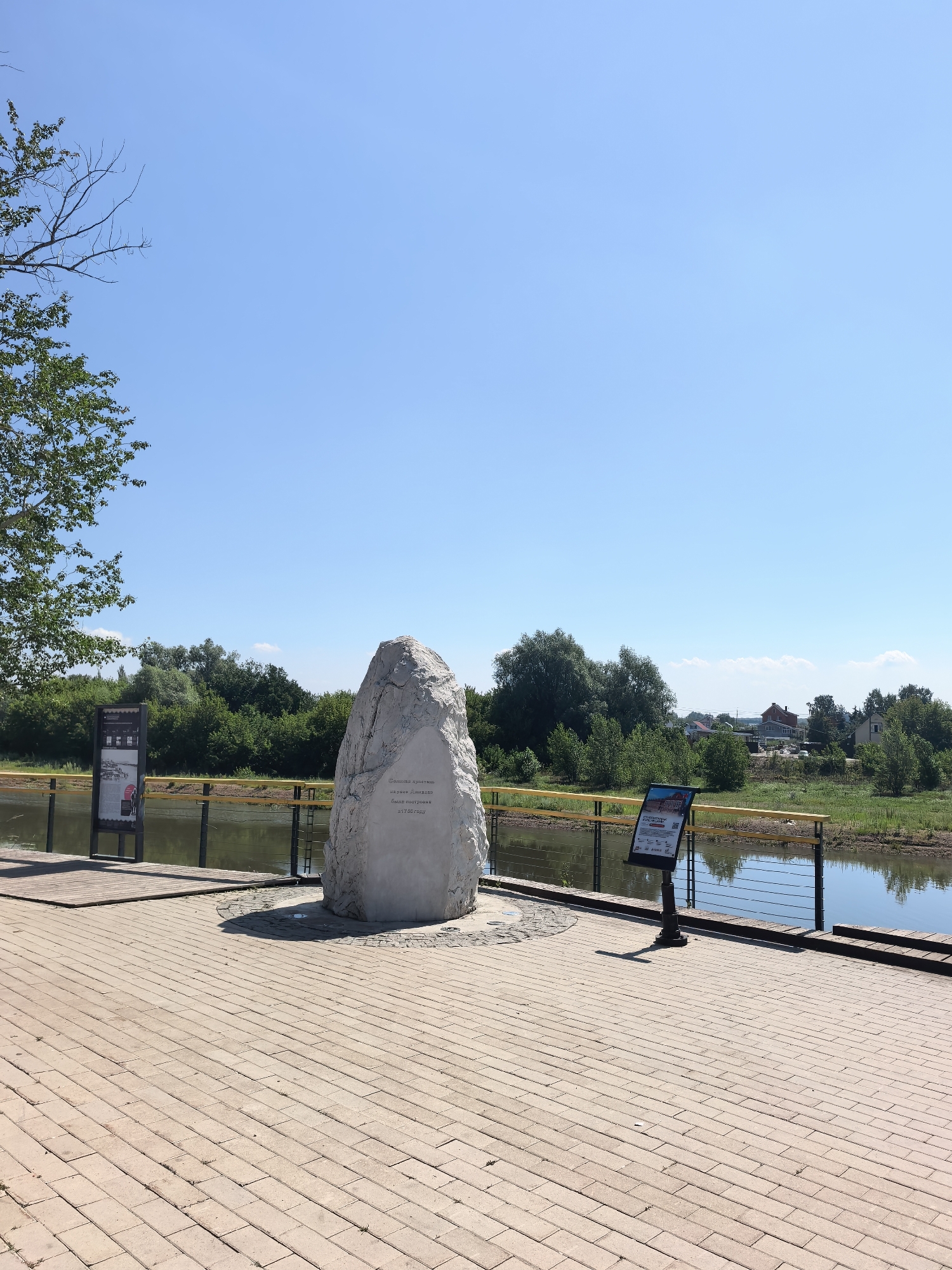 This image depicts a waterside promenade in Sterlitamak, Russia, on a clear, sunny day. In the foreground, a paved walkway made of rectangular light brown bricks stretches across the scene. To the left, a tall, light-colored, rough-textured stone monument stands prominently. The monument is inscribed with text that appears to be in Russian. To the right of the monument, and further into the middle ground, is a black metal post with a sign attached, displaying images and text, also in Russian. A yellow railing runs along the edge of the promenade, separating it from a body of calm, greenish water, likely a river or canal, that flows into the background. Across the water, dense green foliage forms a natural backdrop, with several trees and bushes lining the opposite bank. In the distance, a few houses with red and gray roofs are visible amidst the greenery, suggesting a semi-urban or suburban environment. The sky above is a clear, bright blue with no clouds. The overall scene is tranquil and bathed in natural sunlight.