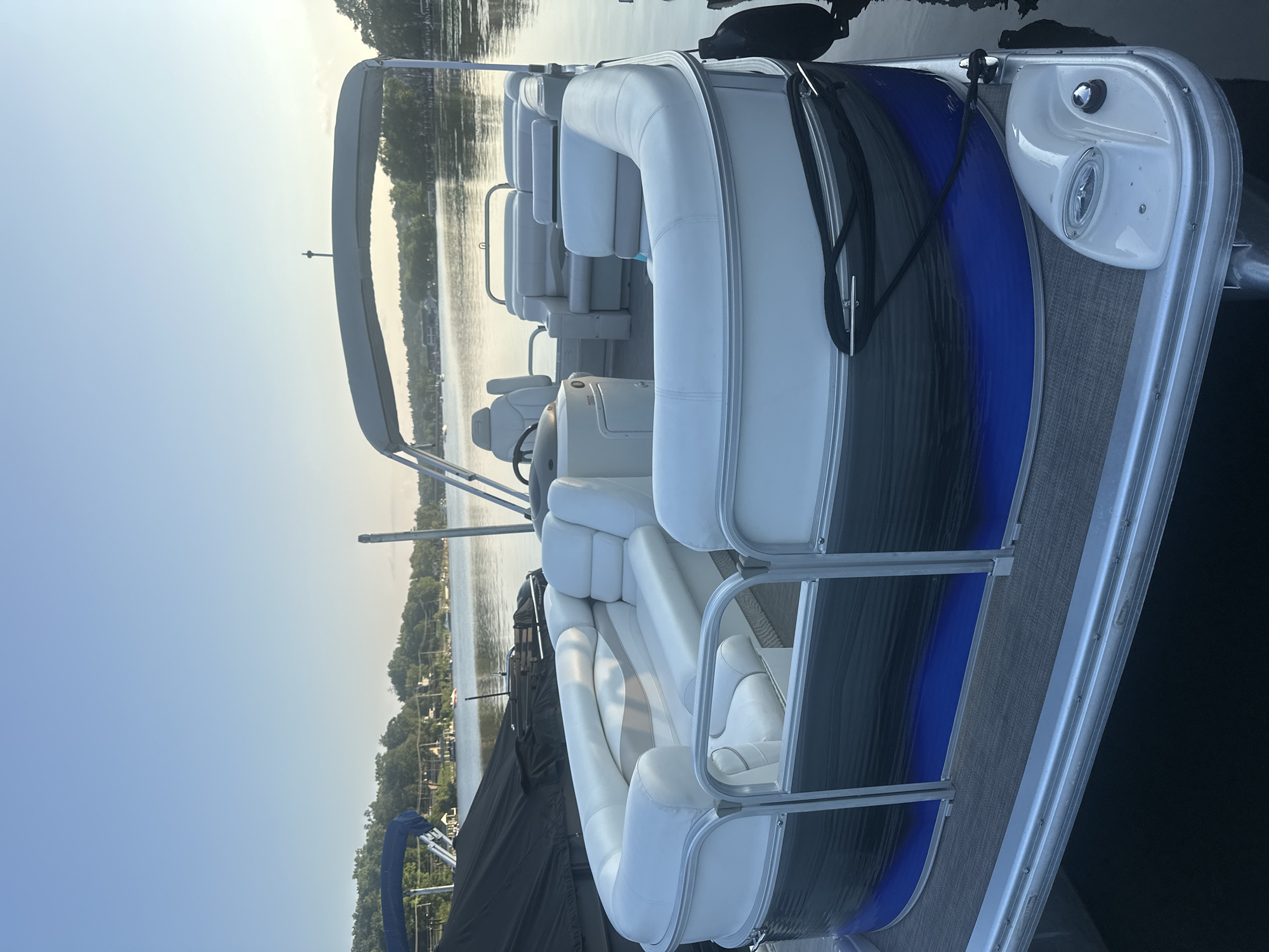 A white and gray pontoon boat is docked on a calm body of water, with a treelined shore and houses visible in the distance. The sky is clear and blue, suggesting it is daytime. The boat has white upholstered seating and a gray canopy. The water appears dark, indicating depth. The scene is peaceful and idyllic.