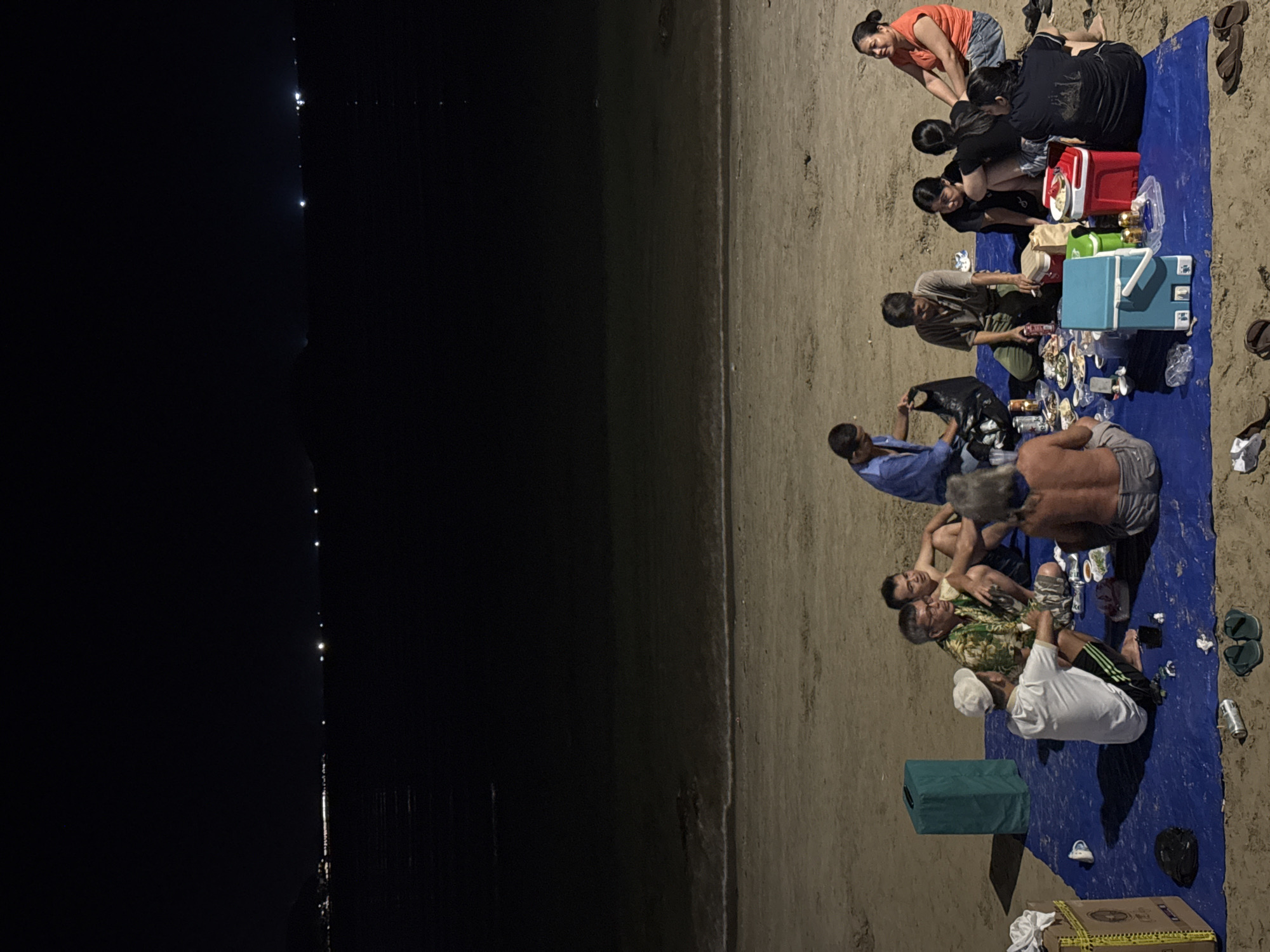A nighttime scene on a beach in Bac Nha Trang Ward, Vietnam, depicts approximately 12 individuals gathered on a blue tarp spread across the sand. The group, composed of both males and females of various ages, is seated or kneeling around an informal setup featuring numerous food containers, plates, plastic cups, and beverage cans, including several beer cans. Two portable coolers, one red and one light blue, are positioned on the tarp. A man in a blue shirt stands near the center-left, holding a black bag, while others are engaged in conversation and interaction. On the sand surrounding the tarp, a teal rectangular object, a cardboard box, and multiple pairs of flip-flops are visible. The background reveals a dark ocean with faint lights on the horizon, indicative of distant boats or coastline activity. The area is illuminated by an unseen artificial light source, creating distinct shadows on the sandy ground.