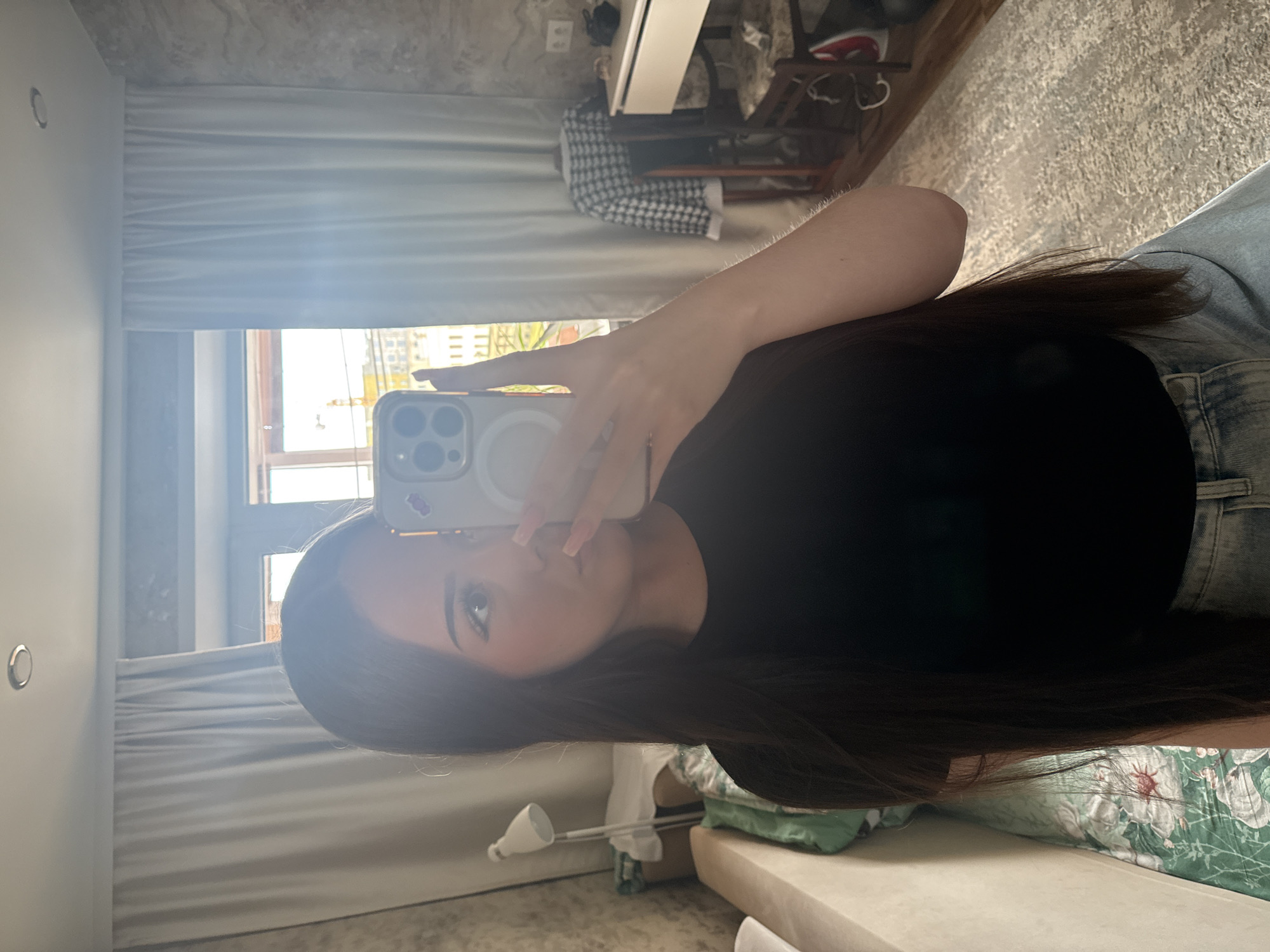 A young woman is captured in a mirror selfie, holding a light-colored smartphone (an iPhone with a MagSafe ring and triple camera system visible) which partially obscures her face. She has long, dark hair and is wearing a black top and light-wash denim jeans. Her reflection occupies the center and lower-left portion of the frame.
The setting is an indoor room, likely a bedroom or living space, in what appears to be an urban environment like Naberezhnye Chelny, Russia. Bright daylight streams in from a large window behind the woman, illuminating the scene and creating a slight glare on the mirror. Through the window, distant city buildings are visible. The window is partially covered by light-colored horizontal blinds or curtains.
In the background, visible in the mirror's reflection, there's a light-colored wall with a small circular fixture. To the right of the woman's arm, a patterned, possibly concrete, ceiling is seen. Below it, a white desk or shelf is present, along with a dark and white checkered garment draped over some furniture. A bed or sofa with green and white floral patterned bedding is also partially visible in the lower right. The floor has a textured, light-colored surface. The overall impression is a casual, everyday moment captured in a brightly lit indoor space. No visible text is legible within the image.