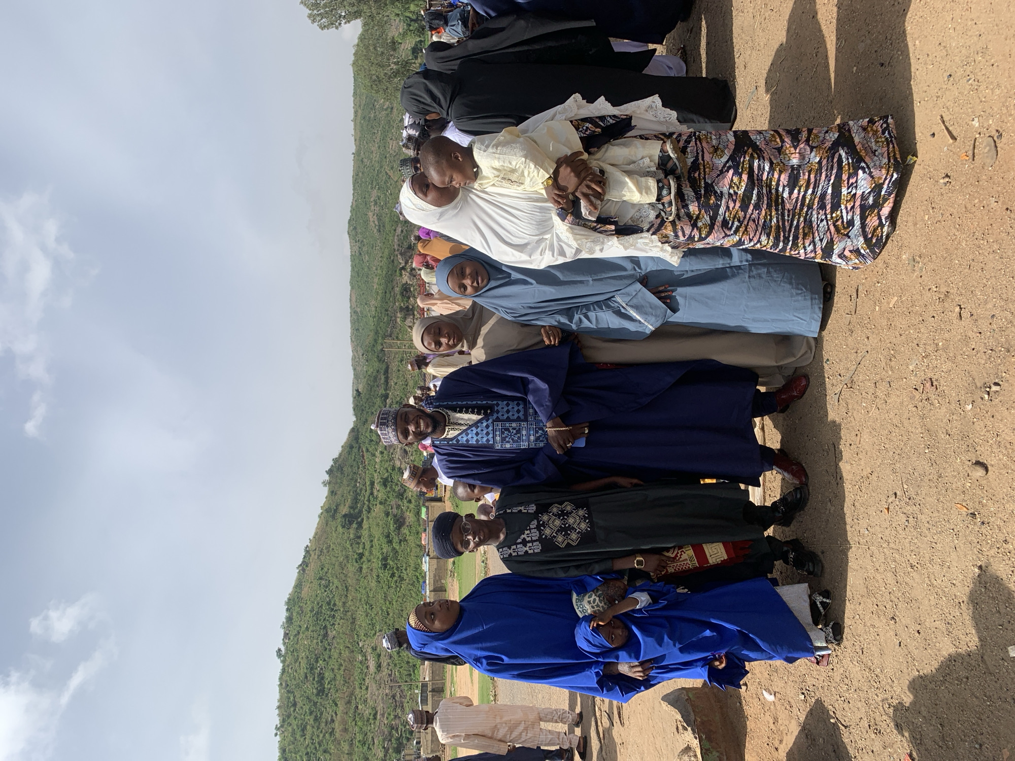 A horizontal, eye-level shot shows a group of about 10 individuals standing in a line on a dirt surface in Naraguta Hausawa, Nigeria. The landscape includes a hill covered with green vegetation and a cloudy sky. The people in the line are dressed in various traditional and religious attires, primarily in colors like blue, white, black, and patterned fabrics. Some are wearing head coverings. At least one person is holding a child. The lighting is bright and sunny, casting shadows on the ground. The overall setting appears to be outdoors, possibly on the edge of the village or town.