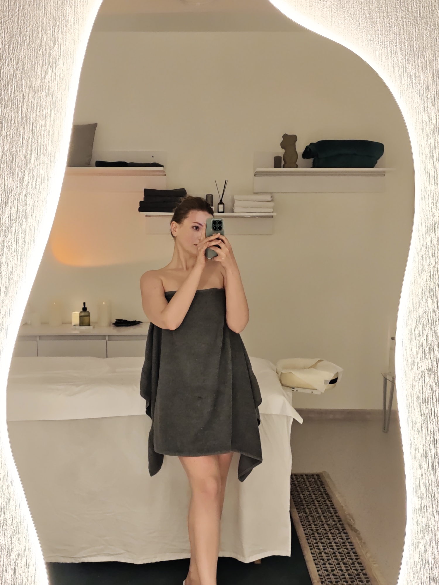 A woman is taking a selfie in a mirror. She is wearing a dark gray towel wrapped around her body. The mirror has an irregular shape with a light border. Behind her is a spa room with a massage table, candles, shelves, and various accessories. The location may be in the city of Vinnytsia, Ukraine.