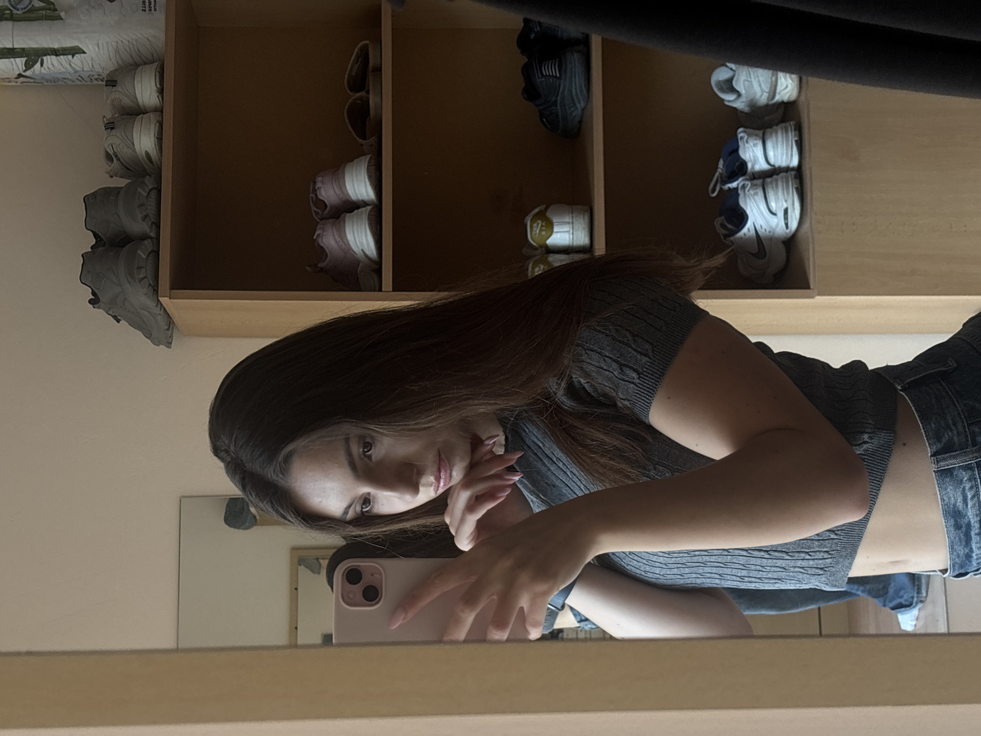 The media file is a portrait of a young woman in Varna, Bulgaria. She is positioned in front of a mirrored surface, angled to capture her reflection and a portion of her surroundings. The woman has long, dark brown hair and is wearing a gray, cable-knit crop top and denim jeans. She is holding a pink smartphone with a dual-lens camera in her right hand, seemingly taking a selfie. Her left arm is bent, with her hand resting on her chin, and she is looking directly at the camera.
In the background, reflected in the mirror and visible in the original image, is a wooden shelving unit filled with shoes. The shelves contain various pairs of sneakers and casual shoes in neutral colors like white, gray, and pink, as well as a pair of black sandals. The overall impression is one of a casual, personal space, likely a bedroom or dressing area. The lighting suggests it is daytime, and there are no external environmental cues indicating specific weather conditions. The woman's expression appears relaxed and self-aware, typical of a casual selfie. No visible text is present in the image.