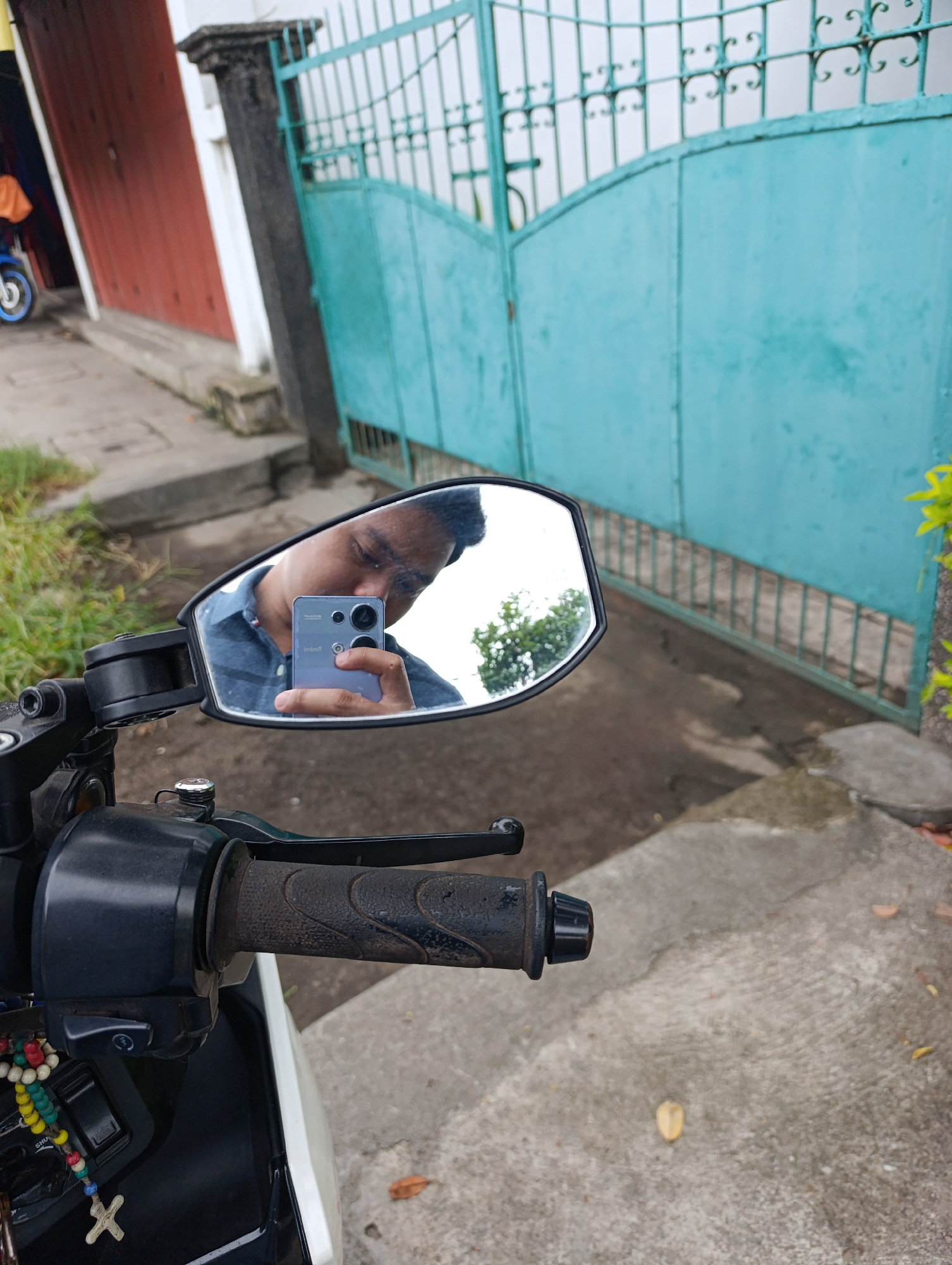 In the city of Unknown, Unknown, an individual is captured in a selfie reflected in a motorcycle's rearview mirror. The person, wearing a blue collared shirt, holds a purple smartphone to take the picture. The mirror also reflects a blurred background of foliage and a turquoise metal gate. In the foreground, the motorcycle's handlebars and a string of colorful prayer beads with a cross charm are visible. The scene is set outdoors on a paved surface, with grass and part of a building with red doors in the background.