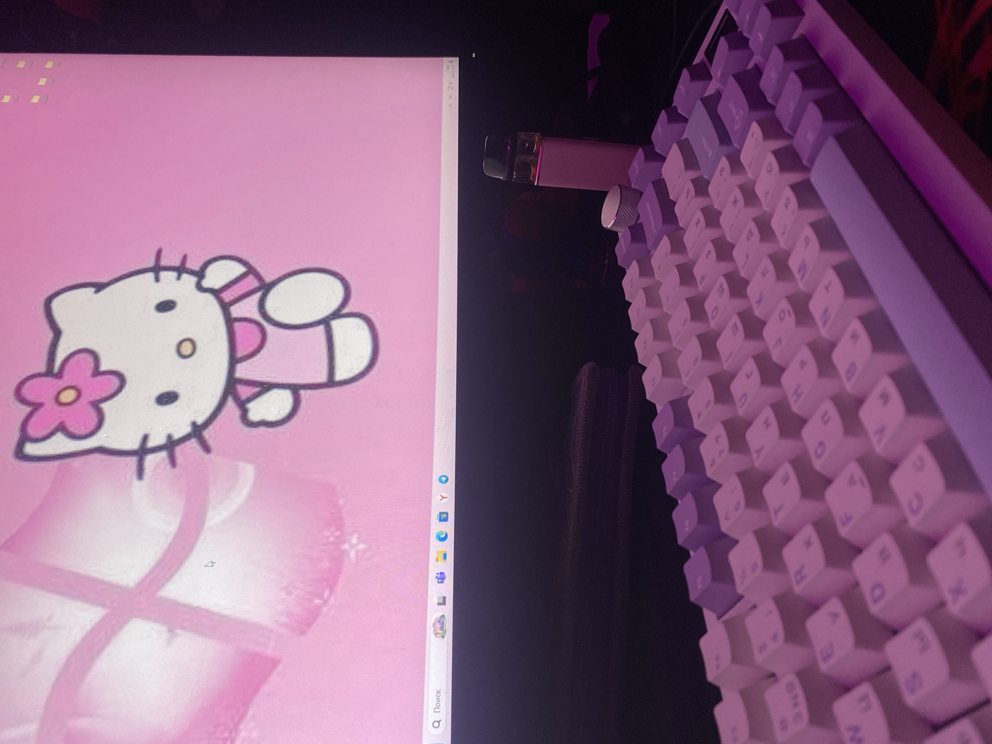 The image shows a portion of a computer monitor displaying a pink Hello Kitty-themed background. To the right of the monitor is a mechanical keyboard with white and light purple keycaps. A pink vaping device sits atop the keyboard. The location and city cannot be determined from the contents of the image.