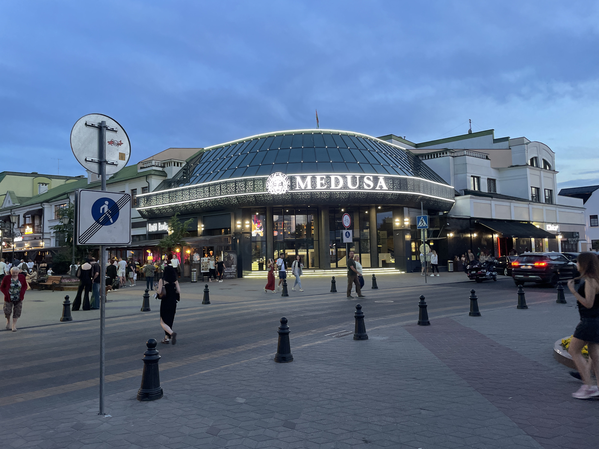 The image depicts a street scene in Brest, Belarus, with the "MEDUSA" establishment as the central focus. The building features a distinctive curved glass roof illuminated by a bright white light strip. The name "MEDUSA" is prominently displayed in large white lettering above the entrance, accompanied by a circular logo featuring a stylized face. Multiple people are visible walking on the street and sidewalk, dressed in casual attire. A black SUV is parked on the street, and a black motorcycle is also present. Traffic bollards line the crosswalk and sidewalk. The sky is overcast and appears to be dusk. A street sign with a pedestrian symbol is visible on the left side of the image.