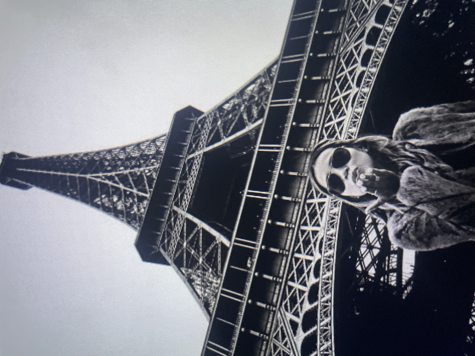The black and white photograph shows a low-angle shot of the Eiffel Tower. In the foreground, a woman wearing sunglasses and a jacket is blowing a kiss towards the camera. The sky is overcast. The image appears to be taken in Paris, France, not Kołobrzeg, Poland.