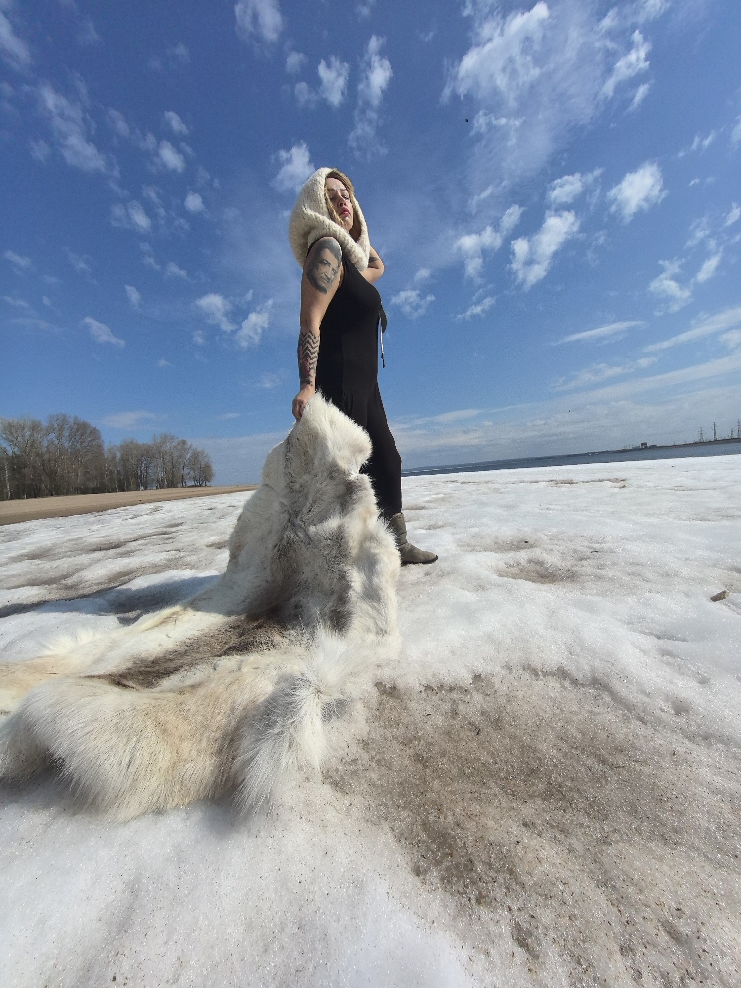 An outdoor, low-angle photograph captures a woman standing on a partially melted, snowy and slushy landscape under a bright blue sky with scattered white clouds. The woman is positioned slightly to the right of the frame, looking upwards and to her right. She wears a black top, dark pants, and light-colored boots, along with a thick, cream-colored knitted hood or cowl. A prominent black and white portrait tattoo of a man is visible on her left upper arm, and other geometric patterned tattoos are on her lower arm.
She holds a large, shaggy white and cream-colored fur hide, possibly a reindeer or similar animal pelt, which trails extensively across the snow-covered ground behind her, creating a striking contrast with the dirty patches of melting snow.
In the background, a line of bare trees borders a sandy or earthy bank, beyond which a wide body of water, likely a river or reservoir, stretches to the horizon. Distant utility poles or industrial structures are faintly visible on the far side of the water. The ground beneath the snow is a mixture of brown earth and shimmering patches of ice and slush, indicating a thawing environment. The clear sky and bright light suggest it is daytime, likely in spring, in a location such as Chaikovsky, Russia. A small, dark, unidentifiable speck, possibly a bird or distant object, is visible in the sky towards the upper right. No explicit text is visible.