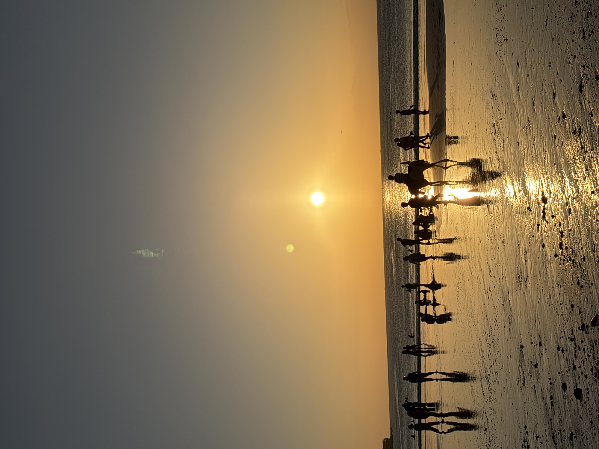 The image captures a sunset scene on a beach in Kuta Selatan, Indonesia. Silhouettes of numerous people are visible on the wet sand, some walking, others standing, and a few engaged in activities like playing or sitting. One prominent silhouette depicts a person riding a horse, with its reflection shimmering on the wet sand. The ocean stretches to the horizon, with gentle waves meeting the shore. The sky is a gradient of warm orange and yellow hues, indicating the setting sun, which is partially visible and casting a bright reflection on the water and sand. The overall atmosphere is serene, with the natural beauty of the sunset dominating the scene.
