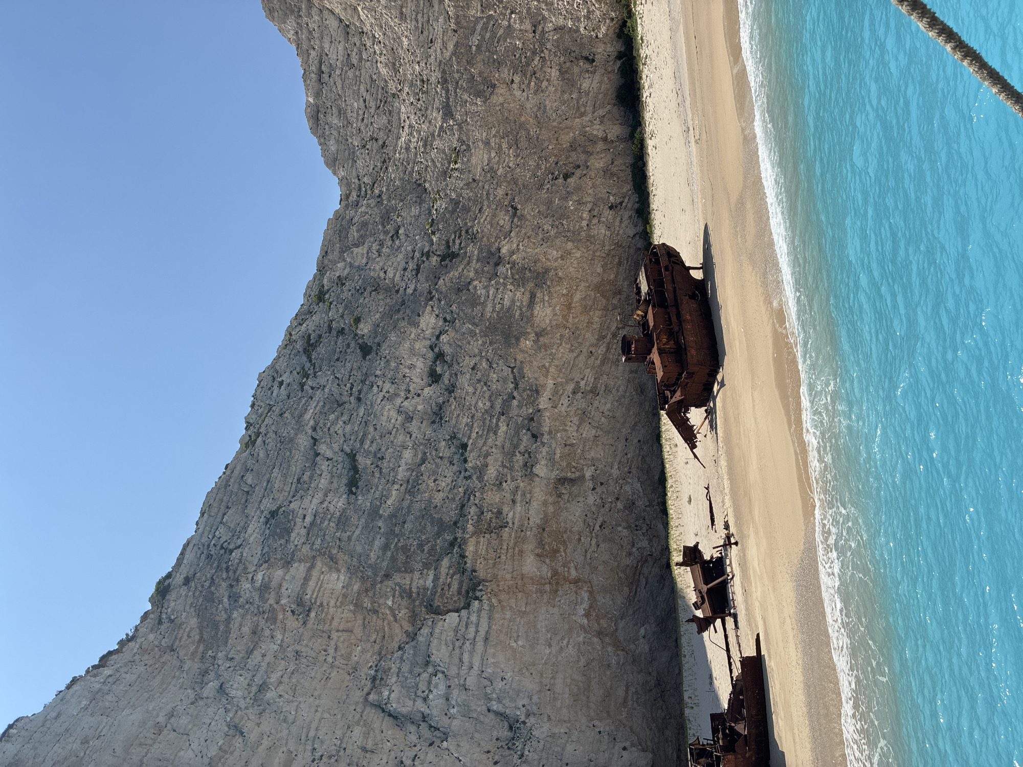 The image depicts the shipwreck of the MV Panagiotis on a sandy beach, likely Navagio Beach, located in the Elaties Municipal Unit, Greece. A steep, stratified limestone cliff rises dramatically behind the beach, its surface textured and showing signs of erosion. The beach itself is composed of light-colored sand, with sparse green vegetation clinging to the base of the cliff.
Several shipwrecks, appearing rusted and heavily decayed, are scattered across the sand. The most prominent wreck is a large vessel with visible hull plating and superstructure, positioned closer to the water. Other, more fragmented wrecks lie further up the beach.
The foreground is dominated by a section of vivid turquoise-blue sea, with gentle waves lapping at the shore, creating white foam. The water appears clear and inviting. A thick rope, possibly from a boat, is visible in the lower right corner of the image, suggesting the photo was taken from a vessel. The sky above is a clear, cloudless blue. The scene is well-lit by sunlight, casting shadows on the sand and highlighting the texture of the cliff face. There are no people visible in the image.