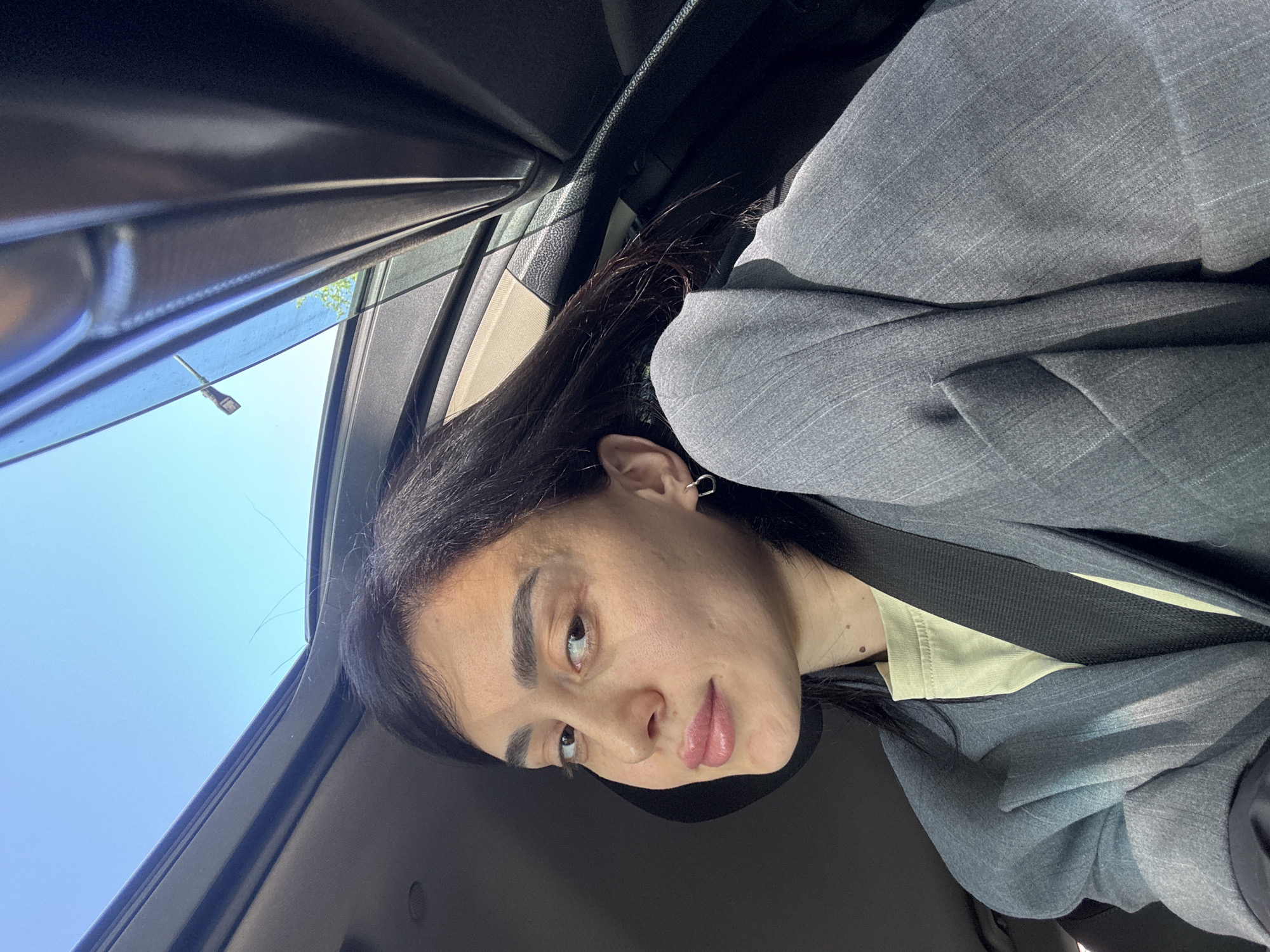 A young person with dark hair is sitting inside a car, wearing a grey blazer and a light yellow shirt. The person is wearing a seatbelt. Outside the car window, the sky is visible and a street lamp. The picture was taken in Tbilisi, Georgia.