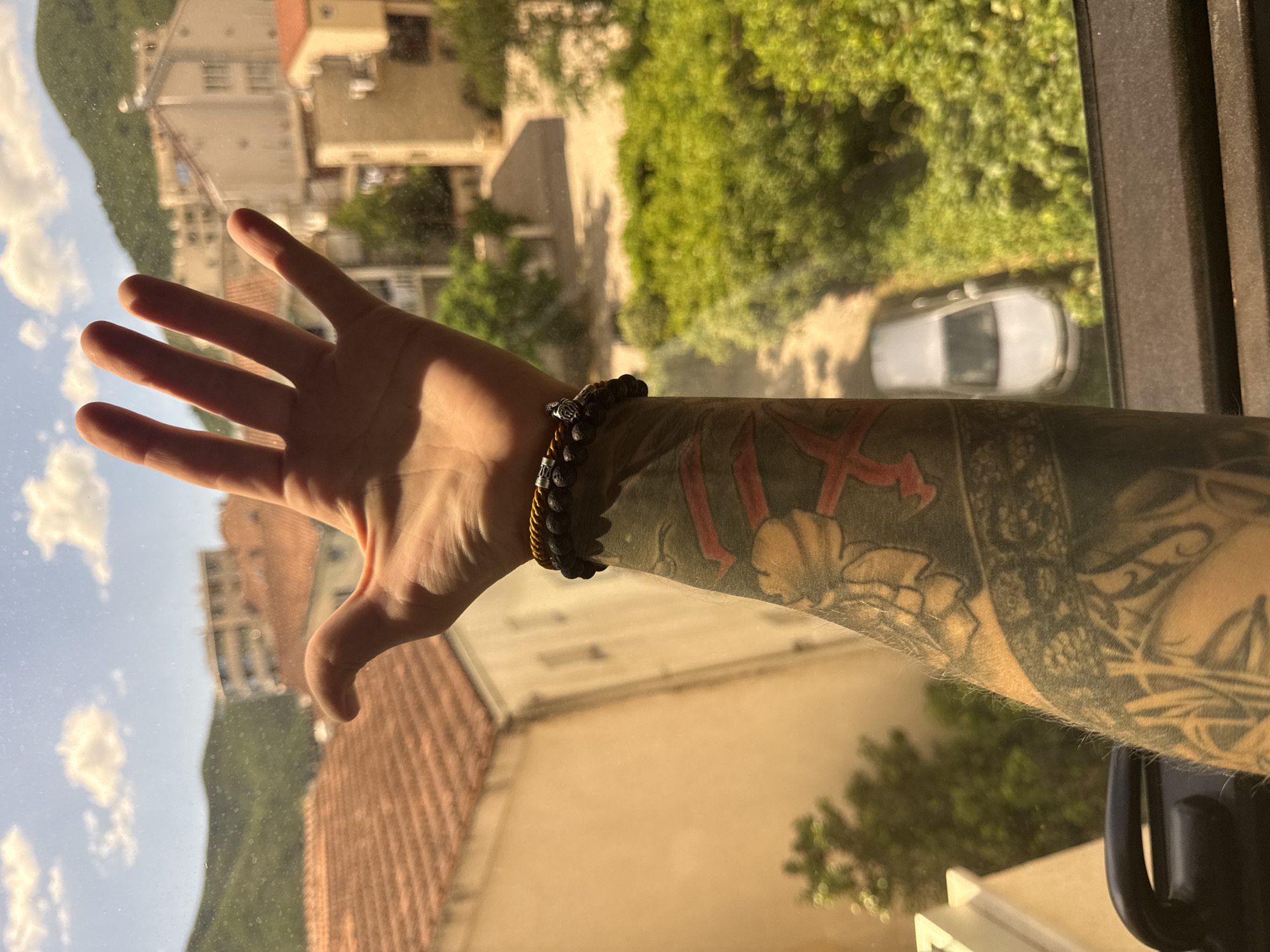 A human arm with extensive tattoos is raised in front of a window, with the hand open and palm facing the camera. The arm is adorned with two bracelets: one with black lava stone beads and a metal charm, and another with yellow and black beads. The background visible through the window depicts a city view, characterized by terracotta tiled roofs, beige buildings, lush green foliage, and a hazy mountain range in the distance. A silver car is parked on a street or driveway, partially obscured by trees. The scene appears to be a residential area within the city of Budva, Montenegro, as indicated by the architectural style of the buildings and the presence of Mediterranean vegetation. The lighting suggests it is daytime, likely mid-morning or afternoon, with sunlight casting shadows and illuminating the scene.