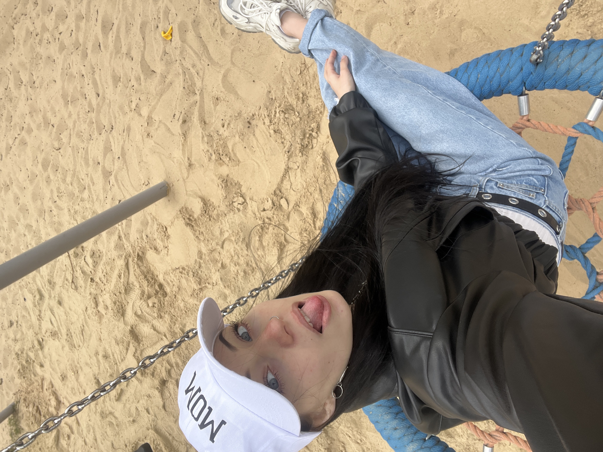 An individual is depicted sitting on a rope swing in a sandy playground. The person, appearing to be a young female, is wearing a white baseball cap with "MOM" in black lettering, a black leather-like jacket, light blue jeans with rolled cuffs, and white and grey athletic sneakers. She has long, dark hair. Her left nostril features a silver hoop piercing, and she wears a silver-toned dangle earring in her left ear. Her eyes are light in color, and her nose appears reddened. The individual is looking towards the camera with her tongue extended.
The swing seat is constructed from woven blue and orange ropes, suspended by metal chains, one of which is visible to the left of the individual. Two grey metal support poles for the swing frame are partially visible in the background. The ground beneath the swing is covered in light tan sand, showing various indentations and footprints. A small, bright yellow, irregularly shaped object lies on the sand in the upper right quadrant of the image, near the base of one of the swing posts. The exact location within Opole, Poland, cannot be determined from the visual information provided; however, the scene is consistent with a public playground.