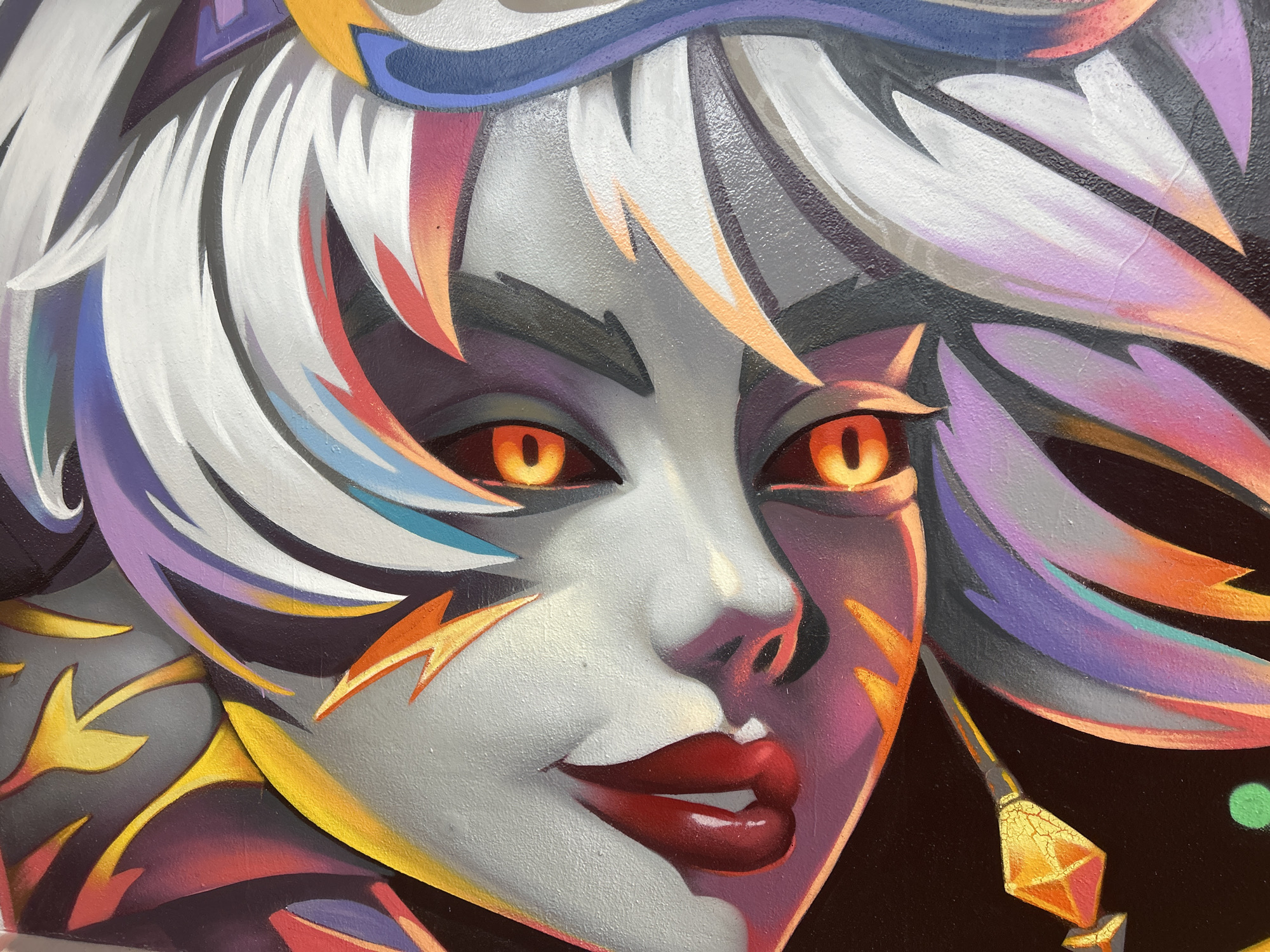 This is a close-up, eye-level photograph of a vibrant mural depicting a stylized, almost anime-inspired female face. The artwork features bold colors and sharp lines, with the subject's skin rendered in shades of gray, highlighted with pink and purple. Her eyes are a striking bright orange with dark pupils, and her lips are a glossy, deep red. Surrounding her face is a cascade of dramatically styled hair in white, purple, pink, and orange, with sharp, angular strokes. Yellow lightning bolt-like shapes adorn her cheeks and forehead. The background is a dark, solid color, with a single green dot visible on the right side. A faceted, golden object with cracked texture is depicted in the lower right portion of the frame. This mural is located on a wall in Sofia, Bulgaria, specifically in the underground passage beneath the Eagles' Bridge (Orlov Most).