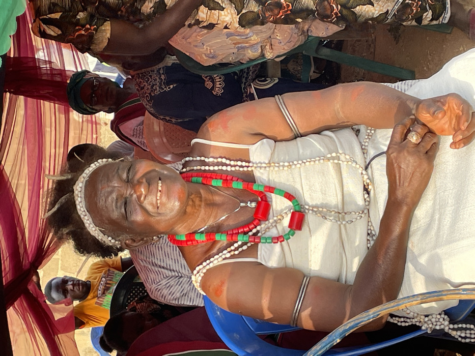The image captures a vibrant moment, likely a celebration or cultural event, featuring an older woman prominently in the foreground, turned slightly to her right and smiling broadly at the viewer. She has dark skin with visible wrinkles around her eyes and mouth, emphasizing her joyful expression. Her teeth are visible in her wide smile.
She is adorned with intricate traditional jewelry and attire. She wears a white, possibly strapless or thin-strapped, top or dress. Around her neck are multiple strands of white beads, which appear to be either small shells (possibly cowrie shells) or pearls, some draped loosely. A striking necklace made of large, alternating red and green cylindrical beads with red spherical connectors is prominently displayed over the white beads. She also wears silver-colored bangles on both upper arms, and an ornate ring is visible on her right hand. Her hair is styled with a white, beaded or shell-embellished headband, and dark, textured hair elements, possibly braids or locs, extend upwards from her head, also appearing to be decorated. Faint reddish-orange markings, suggestive of body paint or ritual markings, are visible on her arms and possibly her face.
In the background, other individuals are partially visible. Behind the main woman's left shoulder, another person, likely a woman, with dark skin, wearing a dark head covering and glasses, looks towards the main subject. Further back on the right, another person in patterned clothing can be seen. In the lower left quadrant of the image, partially obscured by the main woman, a man in a yellow t-shirt with a graphic print and what appears to be white markings on his face or forehead, is also looking towards the left.
The setting appears to be outdoors or under a semi-enclosed temporary structure. Translucent reddish-maroon fabric, resembling netting or sheer cloth, hangs in the background, creating a festive atmosphere, possibly part of a tent or canopy. The ground beneath appears to be dirt or earth. Parts of blue chairs are visible, suggesting seating arrangements for the event. The bright, natural light indicates it is daytime, likely sunny. The overall mood conveyed by the main subject's expression is one of happiness, celebration, and engagement. No legible text is visible in the image.