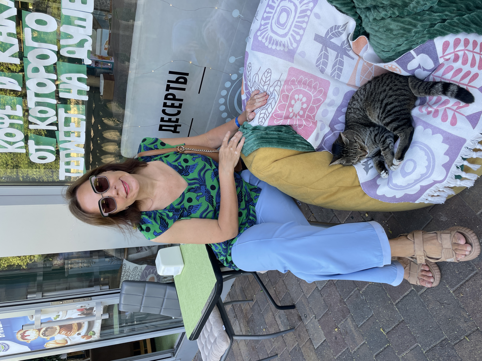 A middle-aged woman with brown hair and sunglasses sits on a colorful cushioned chair in front of a cafe. She is wearing a green and blue patterned top and light blue pants. A tabby cat is sleeping on the cushion next to her. In the background, text in Russian is visible on the cafe window, advertising "coffee and tea, from which it is warm in the soul" and "desserts." The cafe appears to be located on an outdoor seating area with a tiled floor, a small table with a green tablecloth, and a chair with a cushion. The overall impression is one of a relaxed outdoor cafe scene in Sochi, Russia.