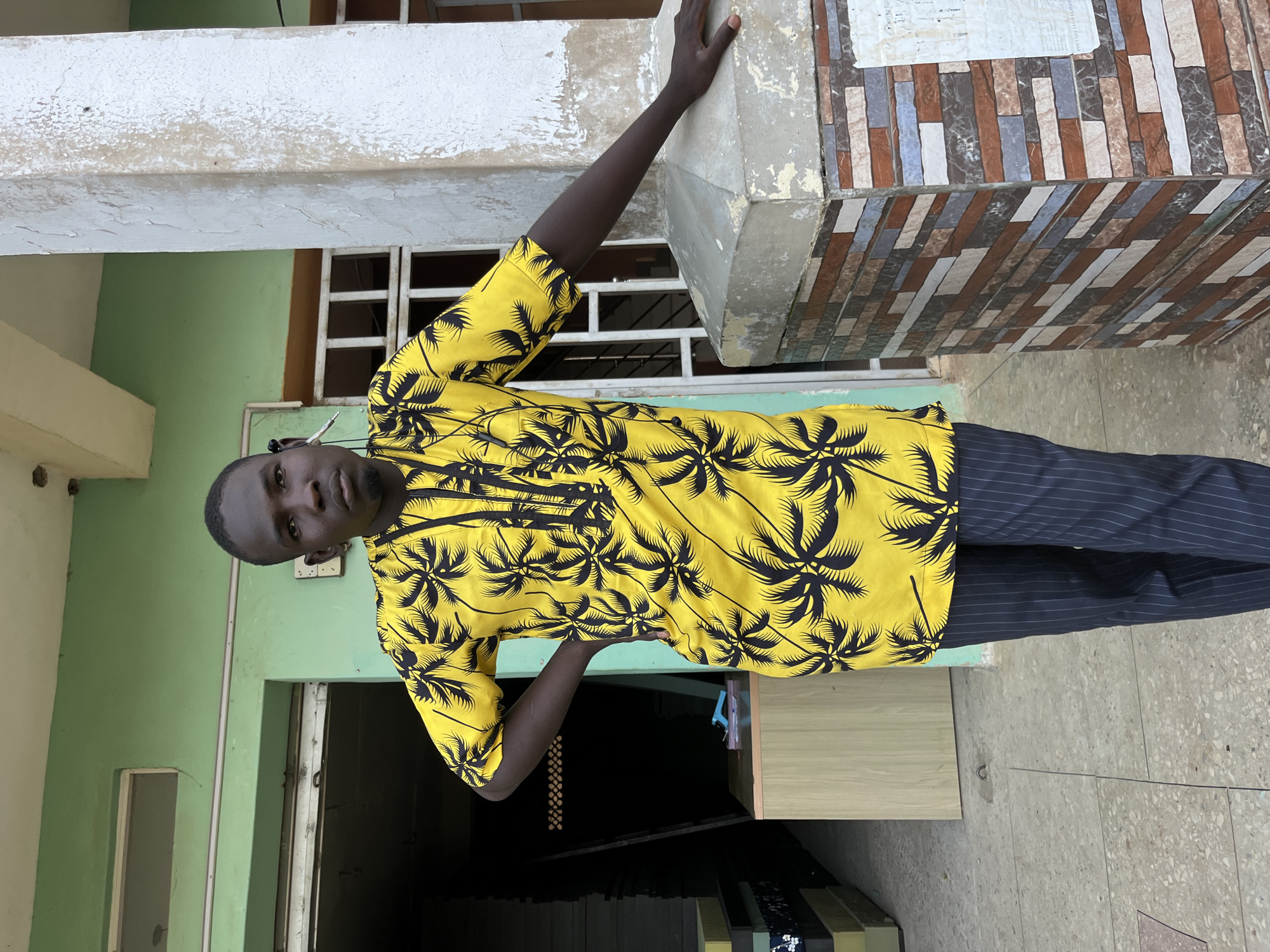 In the city of Gura Top, Nigeria, a young man stands outdoors, leaning against a column. He is wearing a bright yellow shirt adorned with a black palm tree pattern and dark pinstripe pants. He has an earbud in his left ear. The background features a pale green building with a white window frame and a door on the left. To the right of the man, the column is covered in a mosaic of rectangular tiles in shades of brown, blue, white, and gray. The ground is a speckled concrete surface.