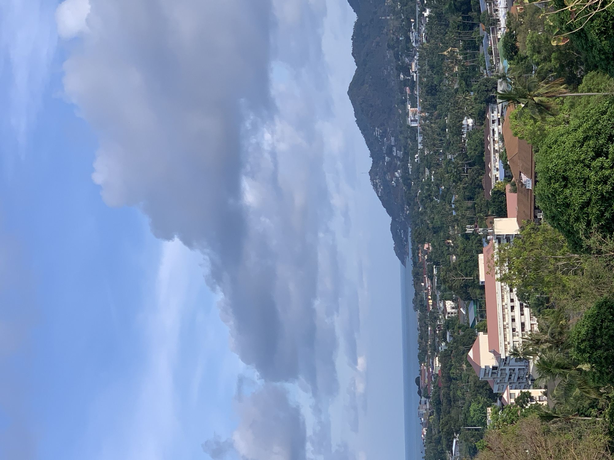 The image shows a coastal landscape in บ้านบางรักษ์, Thailand. The sky is partly cloudy with a mix of light blue and dark gray clouds. A coastline runs diagonally across the center of the image. On the land side, there is a hillside covered in dense tropical vegetation, dotted with buildings and structures of varying sizes, some with reddish-brown roofs. The vegetation consists mainly of trees, including palm trees. The hillside gradually slopes down to the sea. The sea appears calm with a muted blue tone.
