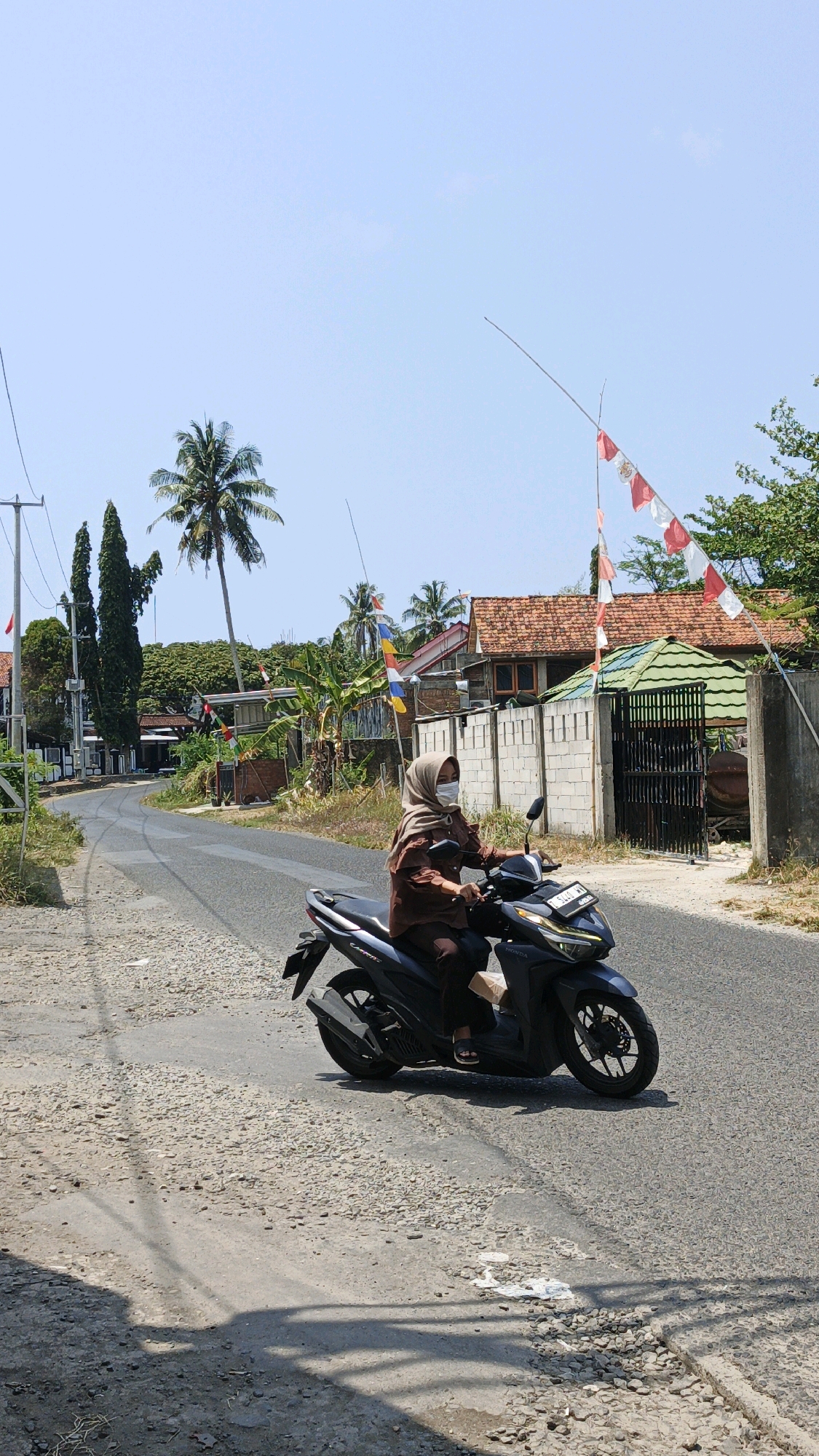 A woman wearing a light brown hijab and a brown long-sleeved shirt is riding a dark gray scooter down a rural road. She is wearing a light-colored face mask. The road is paved but has numerous potholes and is uneven. To the right of the road, there is a wall constructed of light-colored concrete blocks, a black metal gate, and a building with a green roof and reddish-brown tiles. Red and white Indonesian flags are flying on poles to the right of the road, with the flags appearing somewhat tattered. Behind the buildings, there are green trees and palm trees. The sky is clear and blue. The scene is likely set in a village in Indonesia.