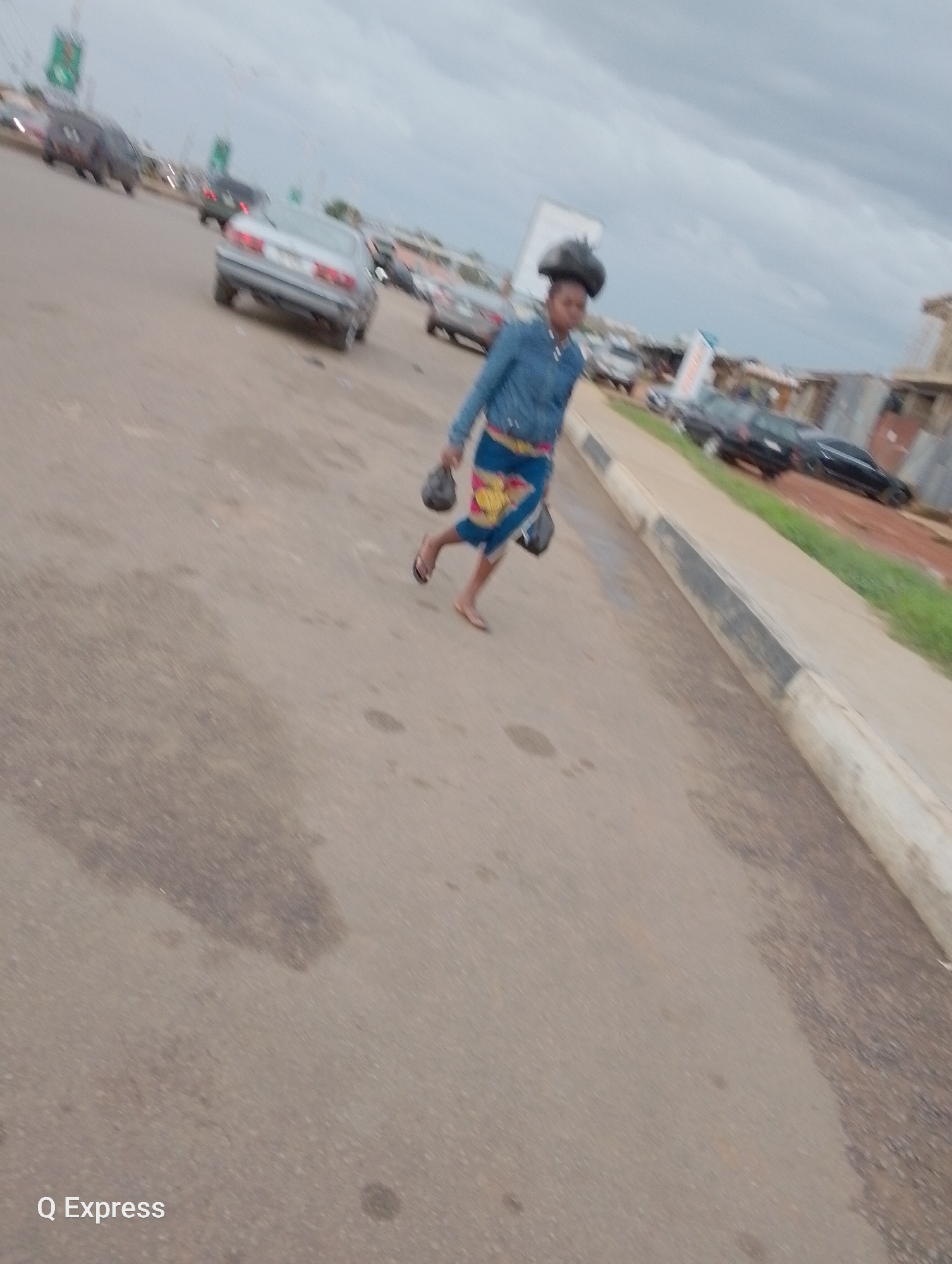 The media file depicts a person walking along a paved road, likely a street in Jos, Nigeria. The individual, appearing to be a woman, is carrying two dark-colored objects, one in each hand, and has another dark object balanced on her head. She is wearing a denim jacket over a colorful, patterned skirt and sandals. The road appears to be a public thoroughfare with several cars visible in the background, suggesting it is daytime. The weather appears overcast, with a cloudy sky. The scene is slightly blurred, possibly due to movement during capture. The presence of buildings and what appears to be a roadside market in the distance further contextualizes the location. The bottom left of the image displays the text "Q Express".