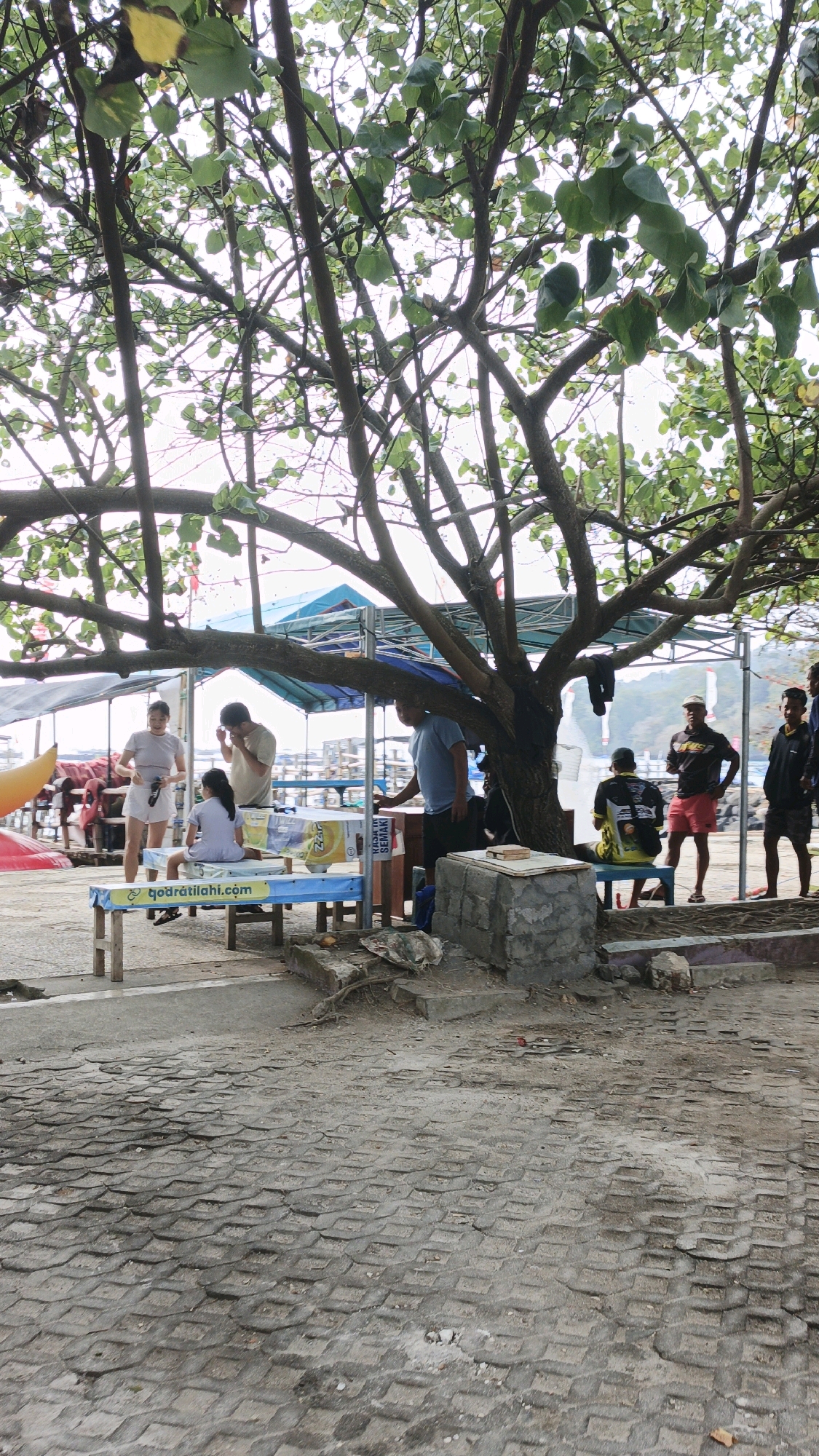 Underneath the branches of a large, leafy tree, a group of people are gathered at a roadside stall near the coast in Desa Pagandaran, Indonesia. The ground in the foreground is paved with interlocking concrete blocks. A blue and yellow-striped table and bench are set up, with a young girl sitting on the bench. To the left, a woman in a light-colored top and white shorts stands near a yellow inflatable object. A man in a light brown shirt is also present near the table, with his hands near his face. Further to the right, a man in a blue shirt stands near a grey stone structure, and another man in a black shirt and red shorts is seated on a bench. Behind them, a blue and teal canopy covers part of the area, and what appears to be a body of water with boats in the distance can be seen. Additional people are visible in the background, standing near tents and structures. The overall scene suggests a casual, outdoor gathering spot, likely a rest area or vendor location near the beach.