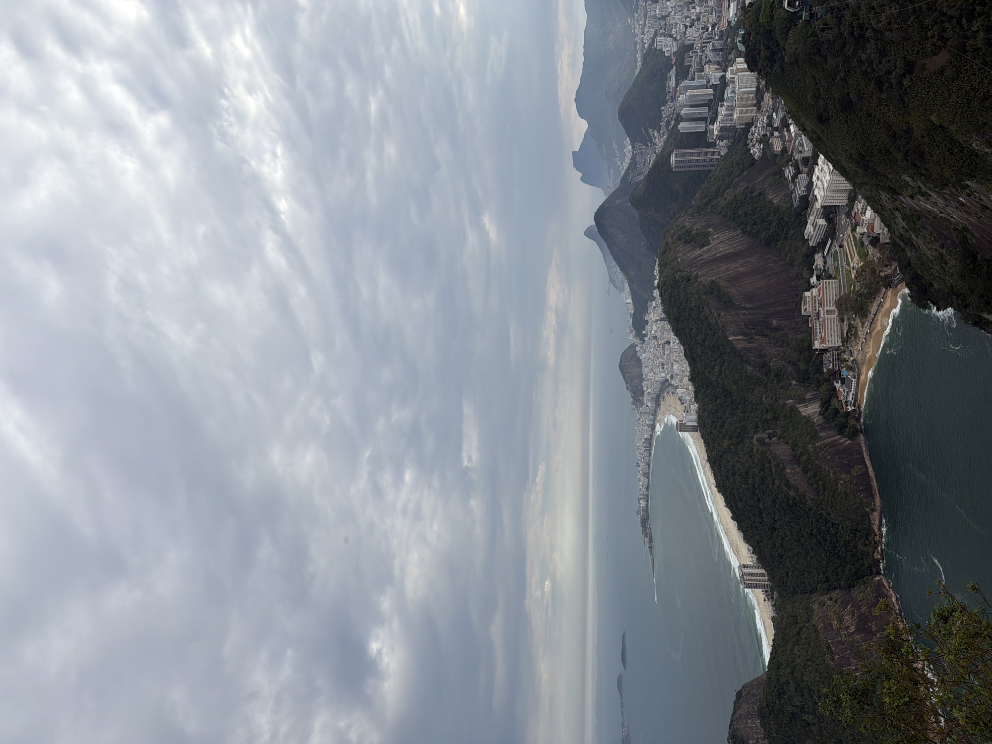 This is an aerial photograph of the city of Rio de Janeiro, Brazil, likely taken from a high vantage point like Sugarloaf Mountain. The image captures a panoramic view of the coastline, with the Atlantic Ocean stretching towards the horizon under a cloudy sky. A crescent-shaped beach, likely Copacabana or Ipanema, is visible, lined with buildings and a sandy shore. Dense green mountains rise from the city, interspersed with urban development, including numerous white buildings and a few taller structures. The water in the foreground is dark and choppy, with some whitecaps indicating waves. The overall scene suggests a coastal city nestled between mountains and the sea, characterized by a mix of natural beauty and urban infrastructure.