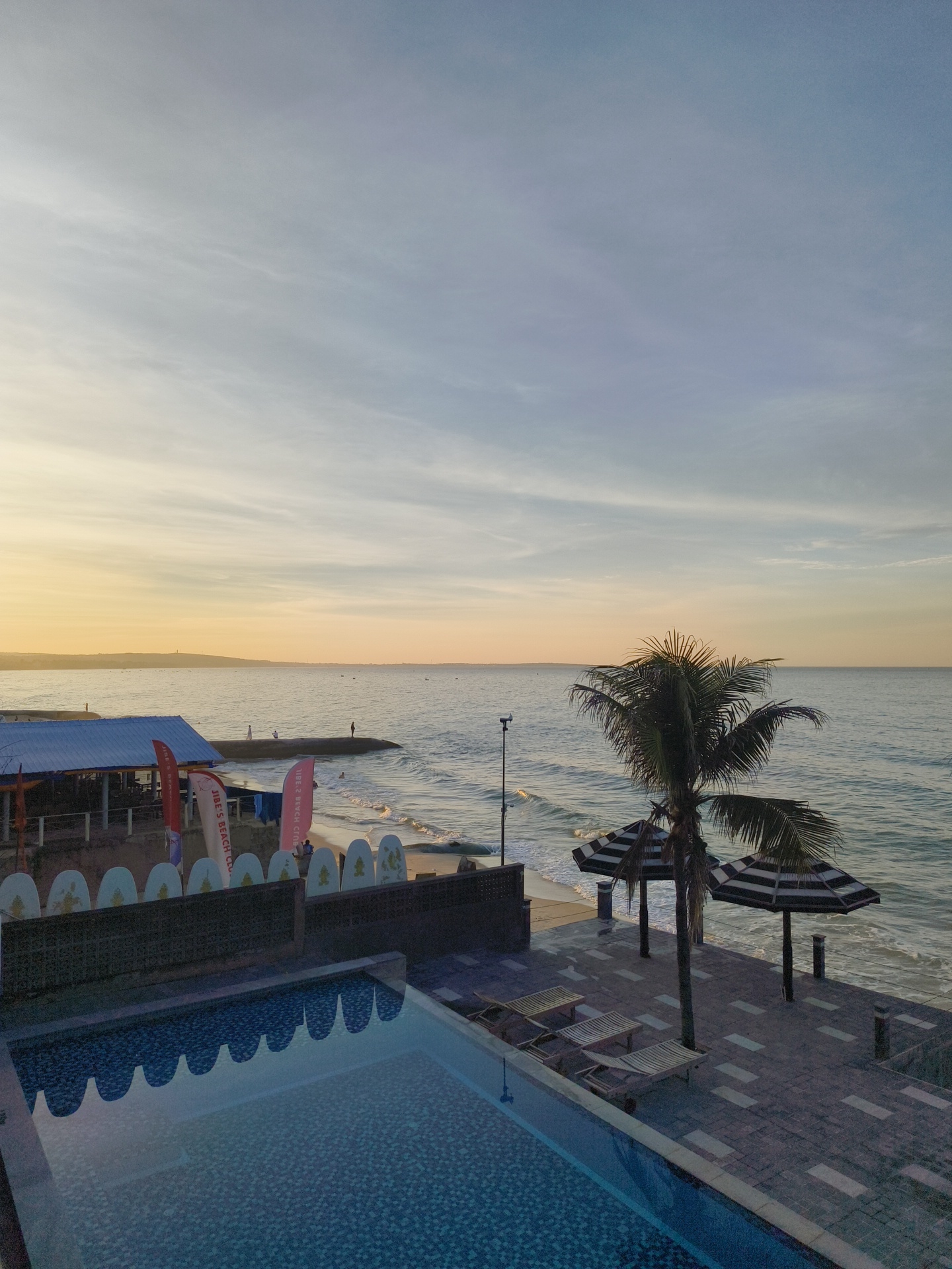 The image depicts a serene coastal scene at dusk, likely in a resort or beach club. In the foreground, a swimming pool with a tiled bottom reflects the sky and surrounding structures. Adjacent to the pool is a paved patio area furnished with lounge chairs and two striped umbrellas, shading the area from the setting sun. A tall palm tree stands near the umbrellas, its fronds spread wide.
Further back, the sandy beach meets the calm ocean, with gentle waves lapping at the shore. A pier extends into the water, with several figures visible, some standing and observing the horizon. A beachside building with a blue roof and open-air seating area is situated behind the beach. Several flags and decorative surfboards line the wall in front of the building. The sky above transitions from a pale yellow and orange near the horizon to a soft blue, with faint clouds scattered across it. The overall ambiance is one of tranquility and relaxation.