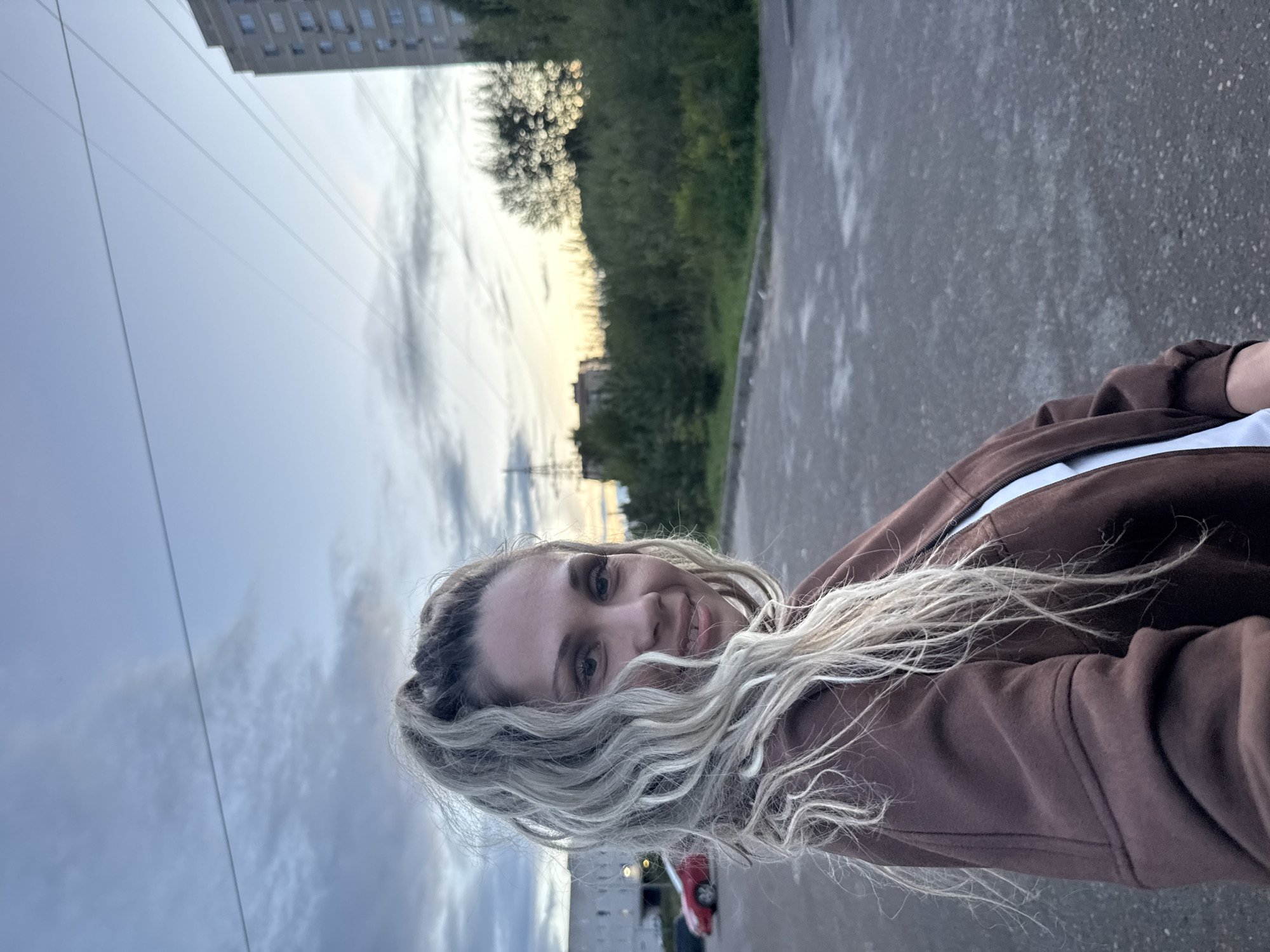 A medium shot captures a Caucasian woman with long, blonde, curly hair pulled into a high ponytail. She is looking directly at the camera with a slight smile. She is wearing a brown zip-up hoodie over a white top. The background appears to be an outdoor urban setting, possibly a parking lot or an open area. In the distance, there is a tall, multi-story building, trees, and power lines against a cloudy twilight sky. A red car is visible in the far left of the image. The scene is static.