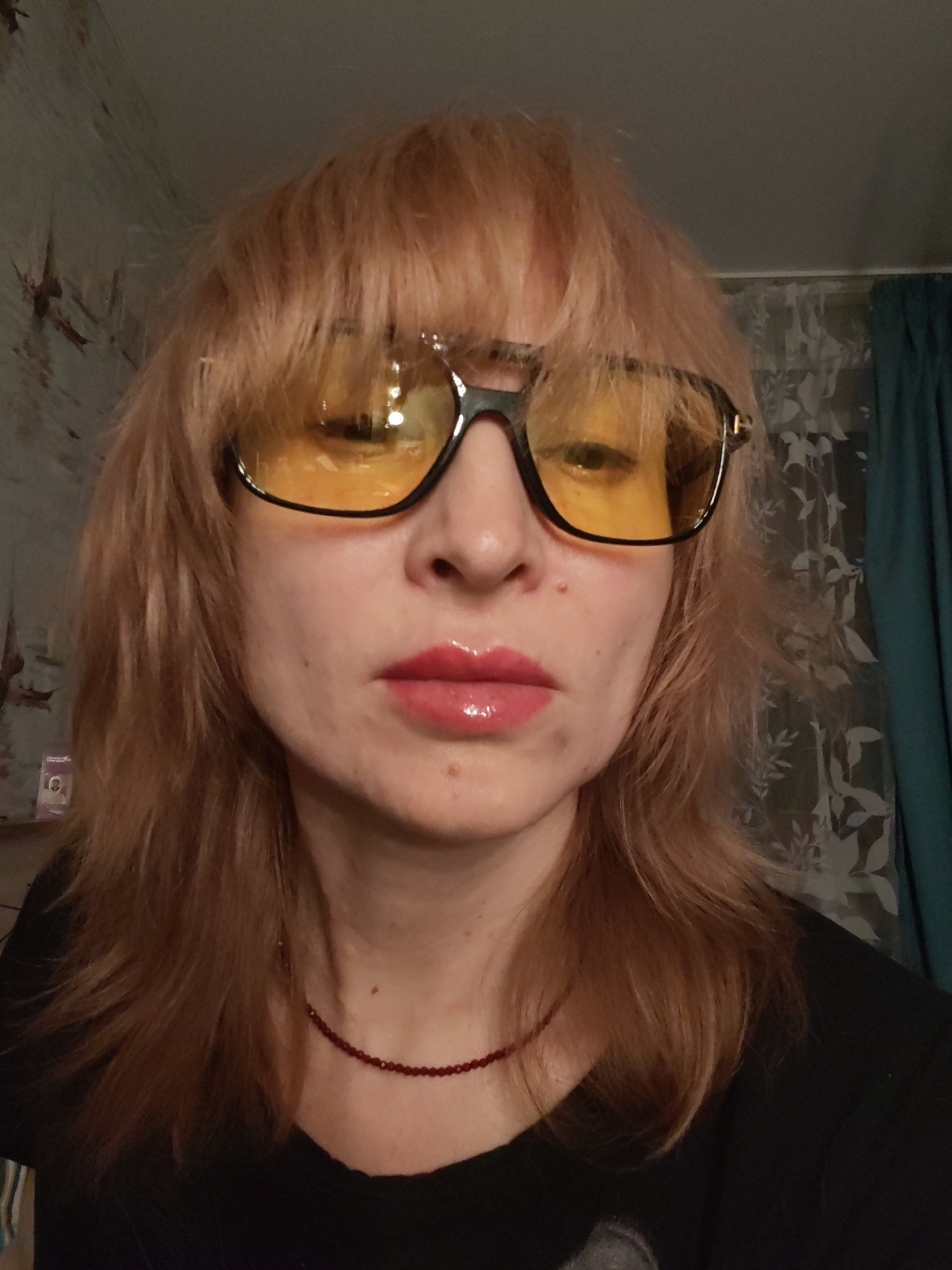 A woman with light brown, layered hair and bangs is wearing large, black-framed glasses with yellow lenses. She has fair skin, pink glossy lips, and is wearing a thin, red beaded necklace. She is wearing a black shirt. The background includes wallpaper with a floral pattern and a dark teal curtain. The location is unknown and information on the city of Chaikovsky is not ascertainable from the image.