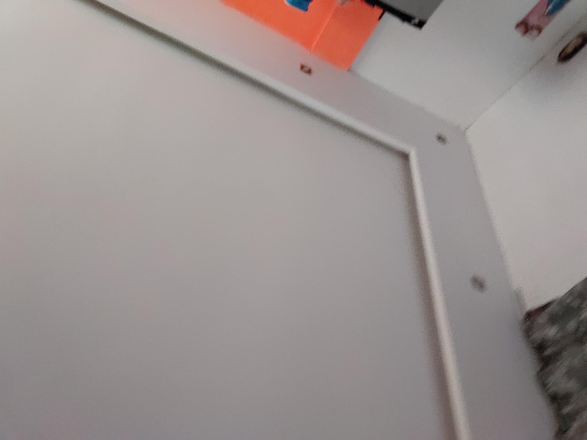 The image presents a close-up, angled view of an indoor setting, dominated by a large, flat, light grey or off-white surface, likely a wall or door panel. This central surface is framed by a lighter, possibly white, trim or molding. Small, dark, circular fasteners, appearing to be screws or bolts, are visible along the trim, securing it to an adjacent surface or to the main panel itself.
In the upper-left quadrant, a sliver of an orange object is visible, possibly paper or cardboard, partially obscured by the frame's edge. Adjacent to it, a small blue shape can be discerned. The upper right corner reveals a corner where two walls or surfaces meet. On the surface extending to the right, faint decorative elements are visible, including a small, partially seen design with pink and blue colors, and a dark, circular or oval object, possibly a light fixture or decoration. The overall impression is of a neutral-toned interior space. No people are visible, nor is any specific activity taking place. There is no visible text. The time of day and weather are not discernible. The image lacks specific cues to confirm the given location context of Santa Ana, Venezuela.