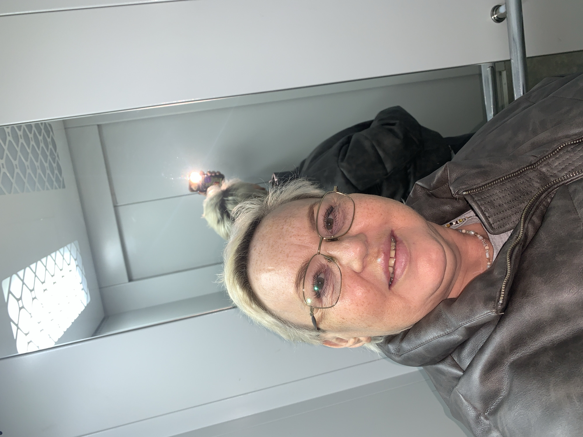 A woman wearing glasses and a brown leather jacket is seen in a selfie, reflected in a mirror. She is smiling slightly. Behind her, in the mirror, part of another black jacket or coat is visible. The setting appears to be a room with white walls, possibly an elevator. A small round object that could be a doorknob is visible on the top part of the wall. The location, Chaikovsky, Russia is not possible to determine from the image.