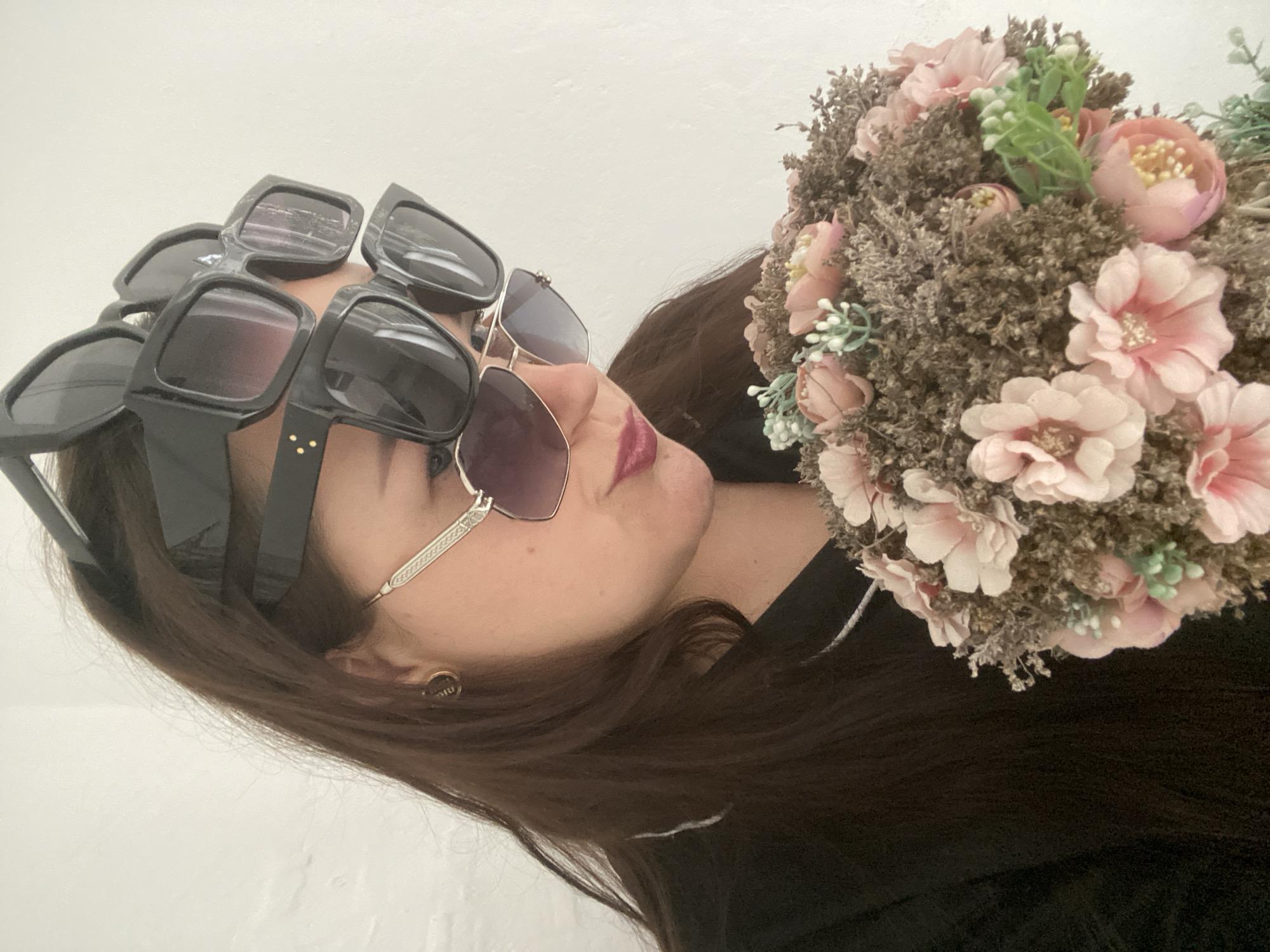 A woman is lying down, wearing multiple pairs of sunglasses stacked on her head. She is holding a bouquet of pink flowers. The background is a white wall. There is no discernible information to indicate this image was taken in Sochi, Russia.