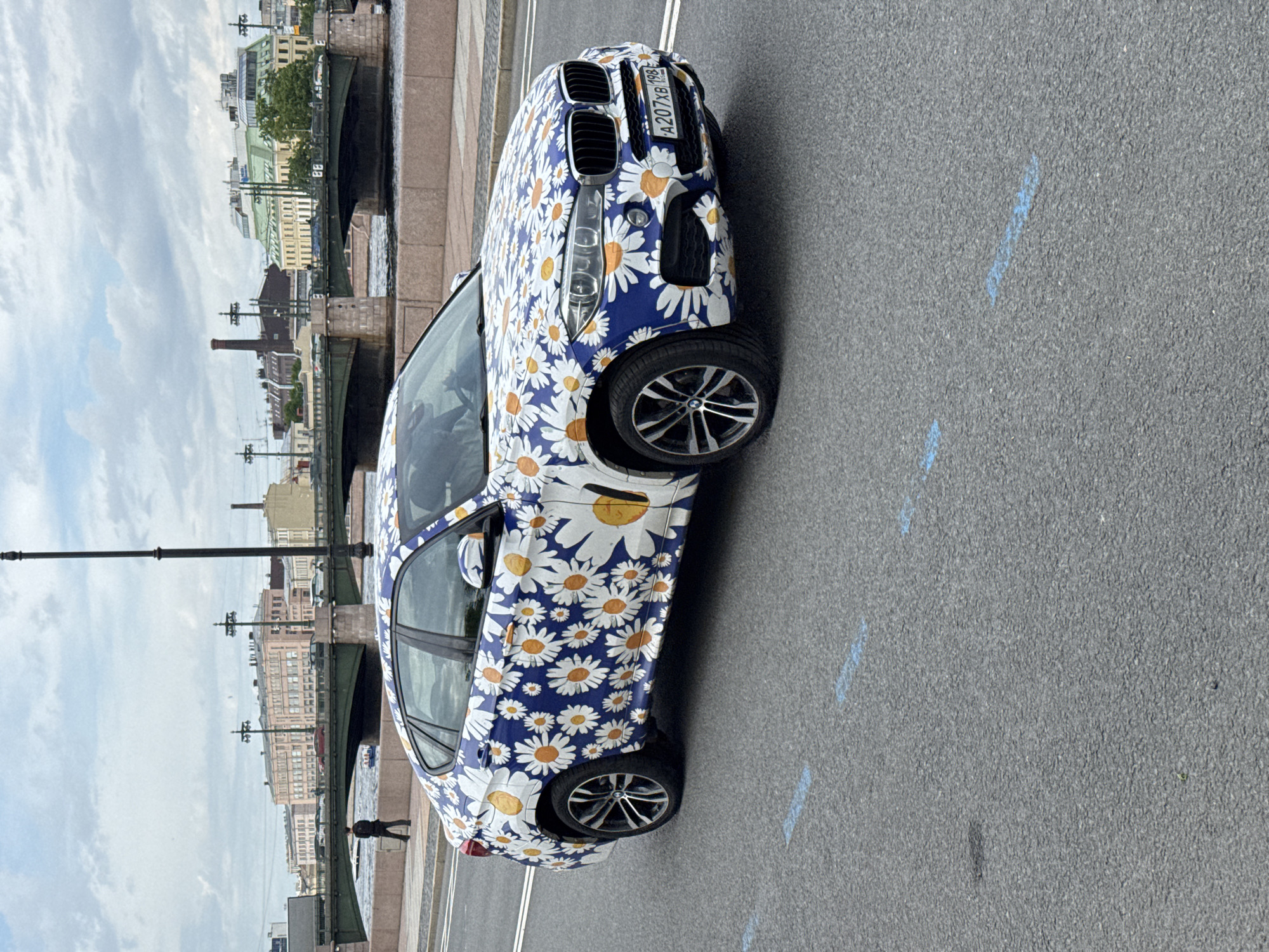 This vertically oriented street scene captures a distinctive blue BMW SUV, adorned with a vibrant white and yellow daisy pattern wrap, parked on an asphalt road. The car's Russian license plate is clearly visible on the front, displaying "A207XB 198."
In the background, a cityscape unfolds under an overcast sky. A sturdy bridge with dark green arched supports spans a body of water, likely a river or canal. Along the far bank, a mix of buildings with varying architectural styles, some featuring light-colored facades and green rooftops, line the horizon. A tall, slender brick chimney rises prominently among the distant buildings. A solitary, small figure of a person can be seen standing near the bridge or riverbank.
The asphalt road in the foreground shows faint, scattered blue markings. The time of day appears to be daytime, with soft, diffused light suggesting an overcast sky. The unique floral design on the car is the most striking visual element, making it highly conspicuous. The scene provides a static snapshot of an urban environment, with the license plate confirming the location as Saint Petersburg, Russia.
