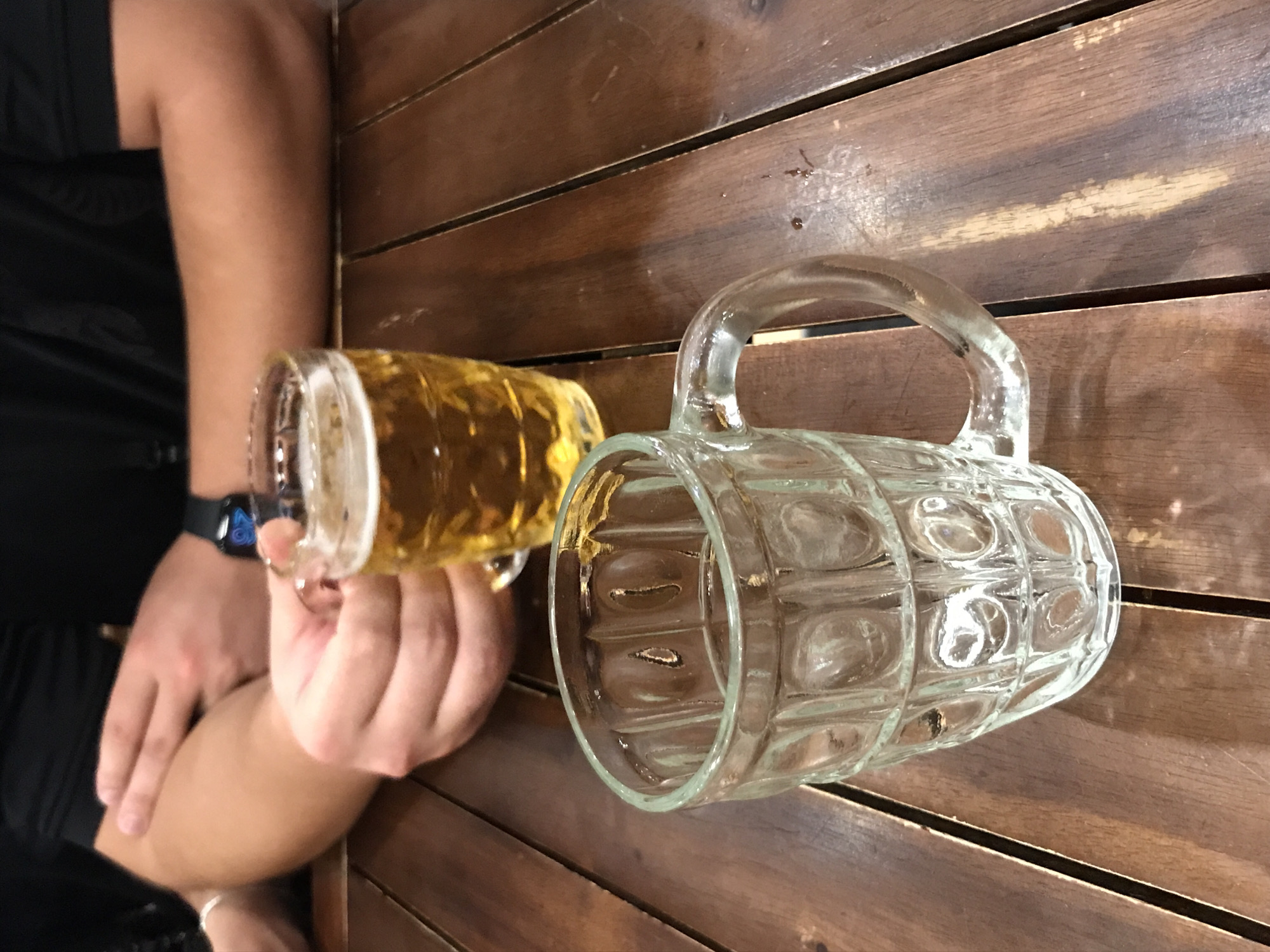 A man holds a glass mug of beer in his left hand. To the right, a similar, empty mug lies on a wooden surface, which appears to be a table with slats. The location is Nha Trang, Vietnam. The man is wearing a black t-shirt and has a watch on his left wrist. The background is blurred, focusing the viewer's attention on the beer and mug.