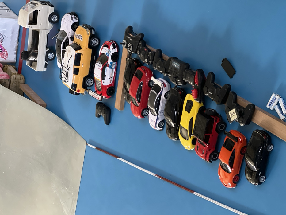 This image depicts an indoor scene, likely a hobby or play area, in Budva, Montenegro. The primary subjects are a collection of twelve remote-control (RC) toy vehicles and at least nine corresponding controllers, arranged on a uniform bright blue surface.
The RC vehicles are predominantly lined diagonally from the upper right to the lower left. From top right to bottom left, the vehicles include: a small white pickup truck, a white SUV, a yellow SUV with a roof rack (resembling a Toyota FJ Cruiser), a red and white race car styled like an Audi R8 with sponsor decals and the number "15," a red sports sedan, a white sports sedan, two black sports sedans, a yellow sports car, a red Jeep-style off-roader, an orange sports car with a rear wing (resembling a Porsche 911), and a black sports coupe.
Most of the black RC controllers are aligned on a narrow wooden plank that runs parallel to the line of vehicles. One controller features a white label. Additionally, three white AA batteries and a small black rectangular object (possibly a battery cover) are visible at the bottom right.
In the upper left portion of the frame, a large, reflective silver metal sheet forms an inclined ramp, supported by small wooden blocks. A thin, red and white striped line extends diagonally from the lower left towards the center, bordering the blue surface. Blurred elements in the background, including a cardboard structure, suggest further elements of a track or diorama. No people are present in the scene.