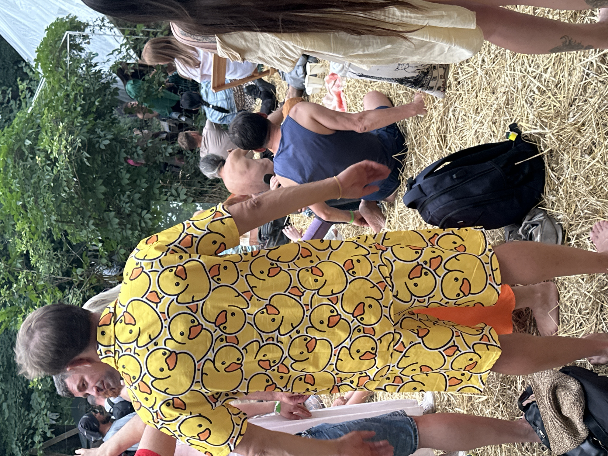 In Kyiv, Ukraine, amidst a grassy, outdoor setting with lush green foliage and a white tent structure, a man in a vibrant yellow shirt adorned with a repeated duck pattern stands prominently in the foreground. He is viewed from behind, showcasing the full print of his attire. Around him, other individuals are seated or standing on a ground covered with dry straw. A man in a blue tank top is kneeling, and another person in a tan shirt is shirtless, their backs turned to the camera. A black backpack rests on the straw near the kneeling man. The atmosphere suggests a casual gathering or event.