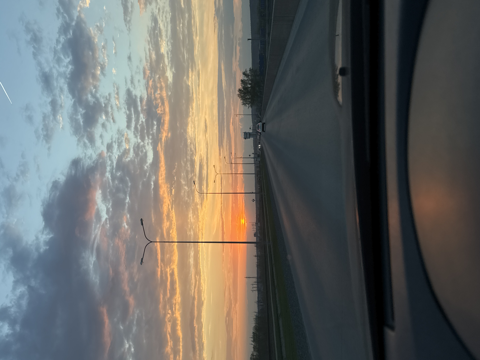 The image is taken from inside a vehicle moving on a road in what is likely the city of Chaikovsky, Russia, based on the prompt. The sky is filled with clouds painted in hues of orange and blue from the sunset. Street lights line the side of the road. There is a power plant/industrial facility along the side of the road. There is a single car ahead on the road. The interior of the car is visible on the right.